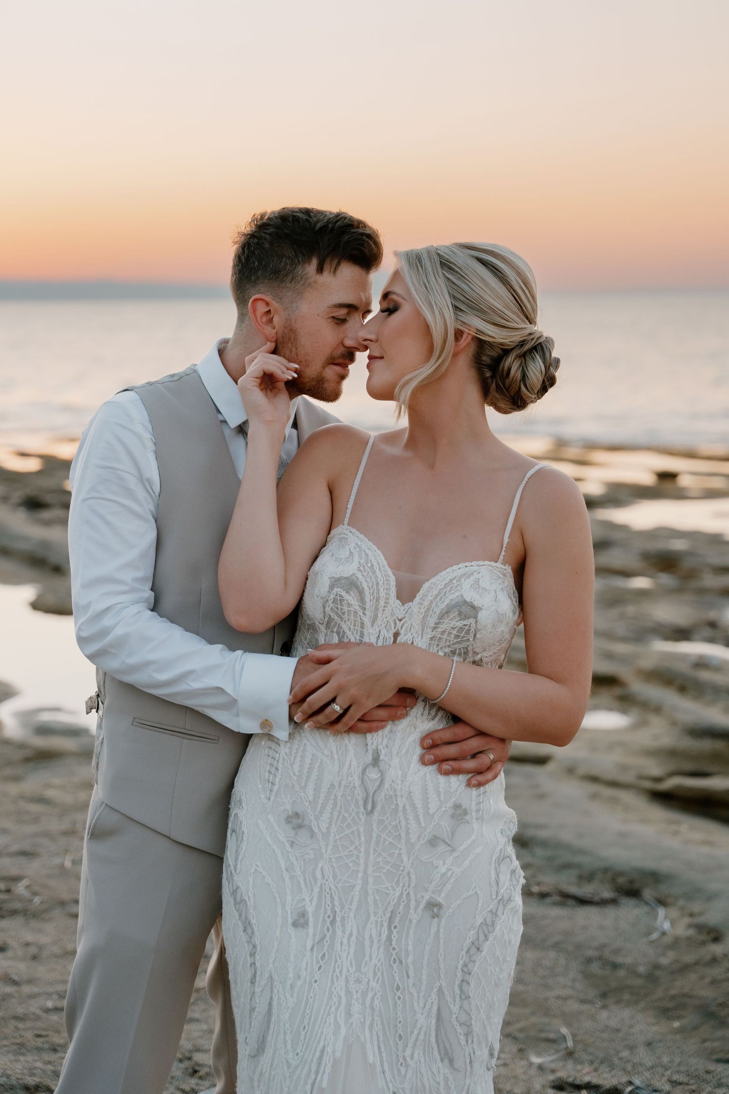 Romantic beach portraits at sunset show tender moments between newlyweds against rocky coastal backdrop.
