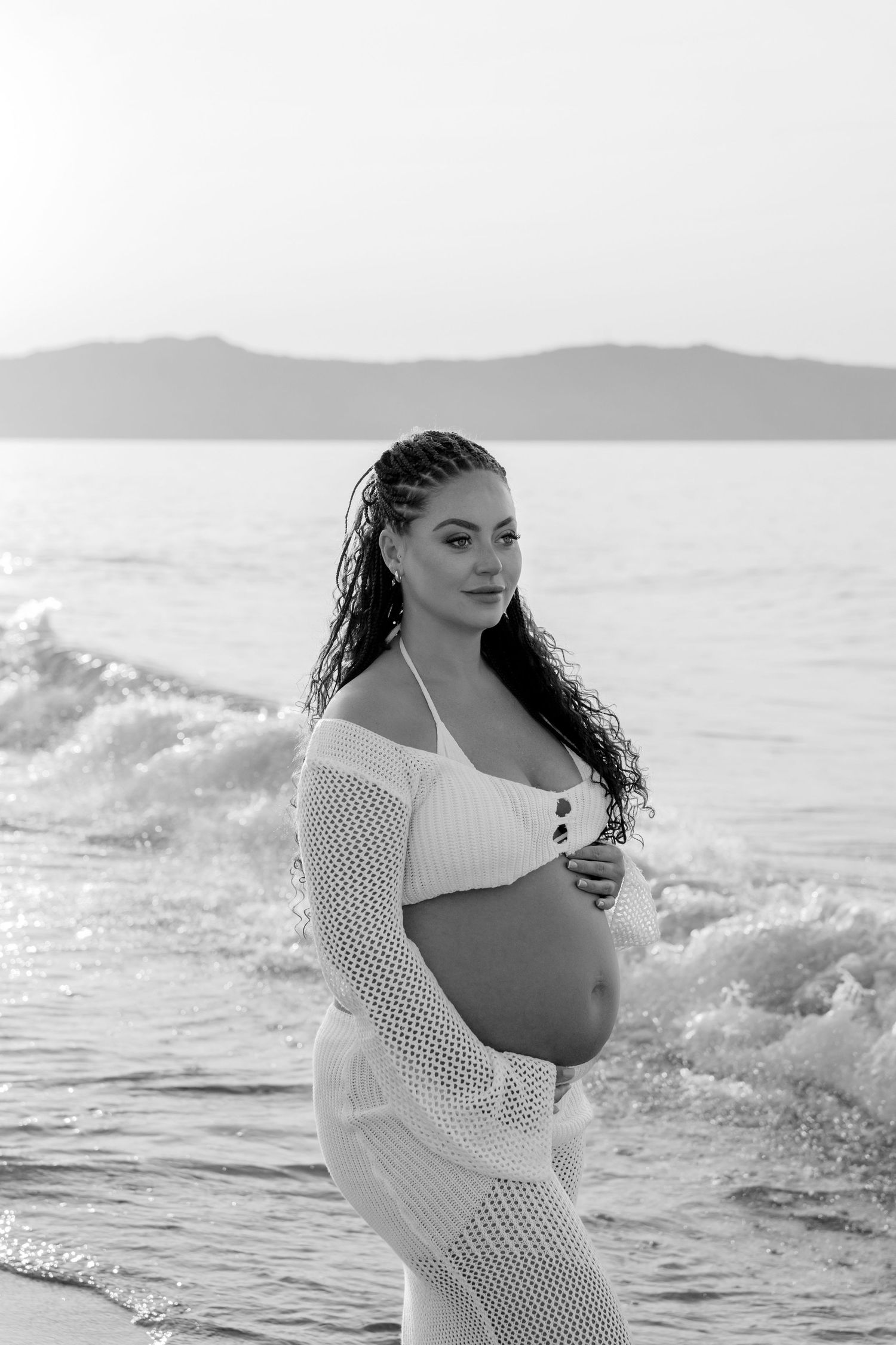 Maternity photo shoot at the beach showing an expectant mother in white mesh coverup against ocean waves at sunset.