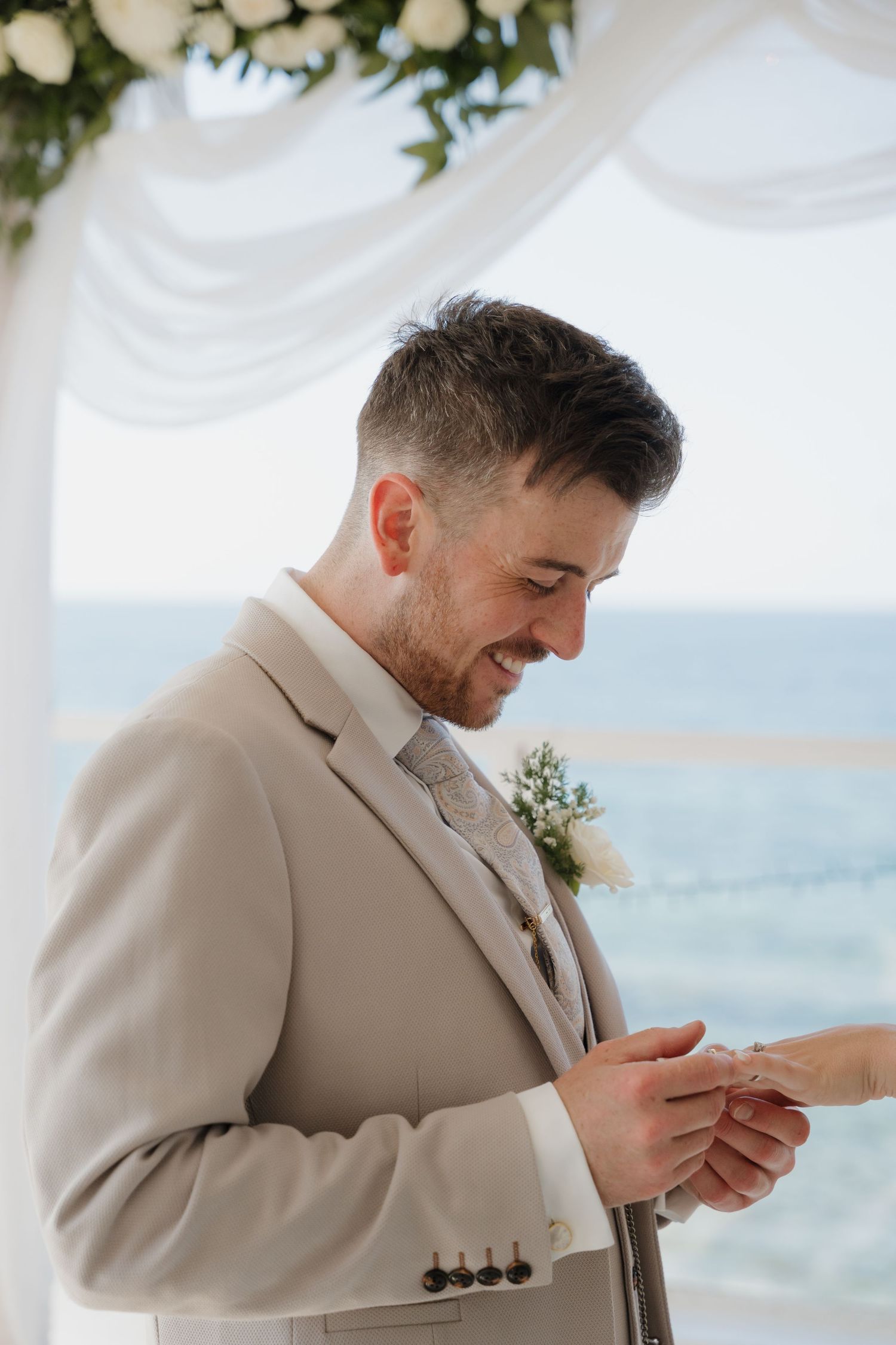 A groom in a light beige suit smiles while placing a ring during a beachside wedding ceremony.