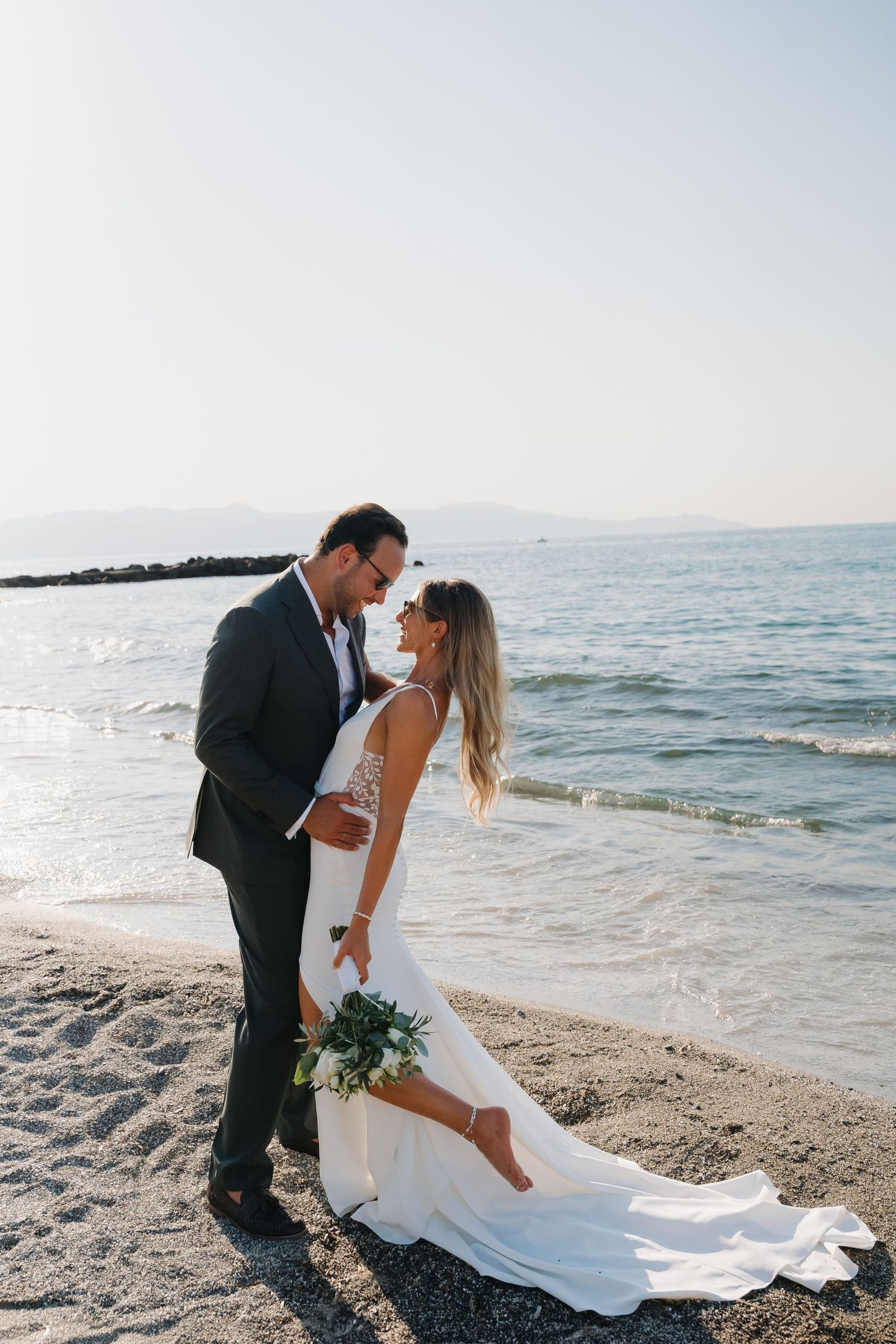 Newlyweds embrace on a scenic beach with flowing white wedding dress and ocean backdrop.