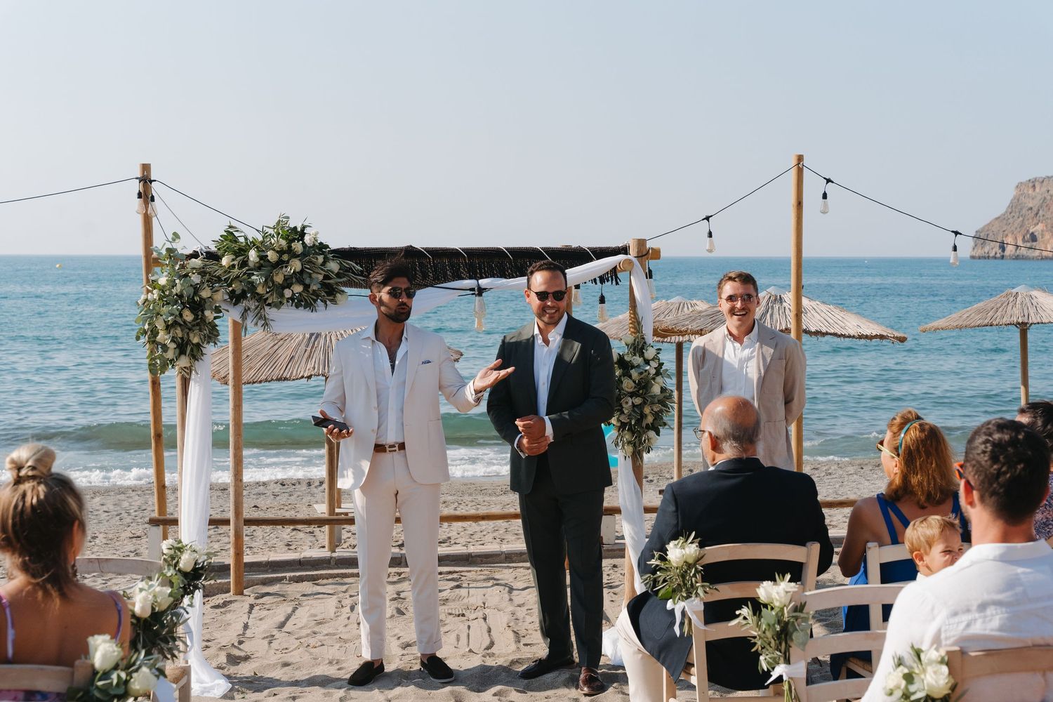 A beachside wedding ceremony takes place under a rustic wooden arch with the ocean and blue sky as backdrop.