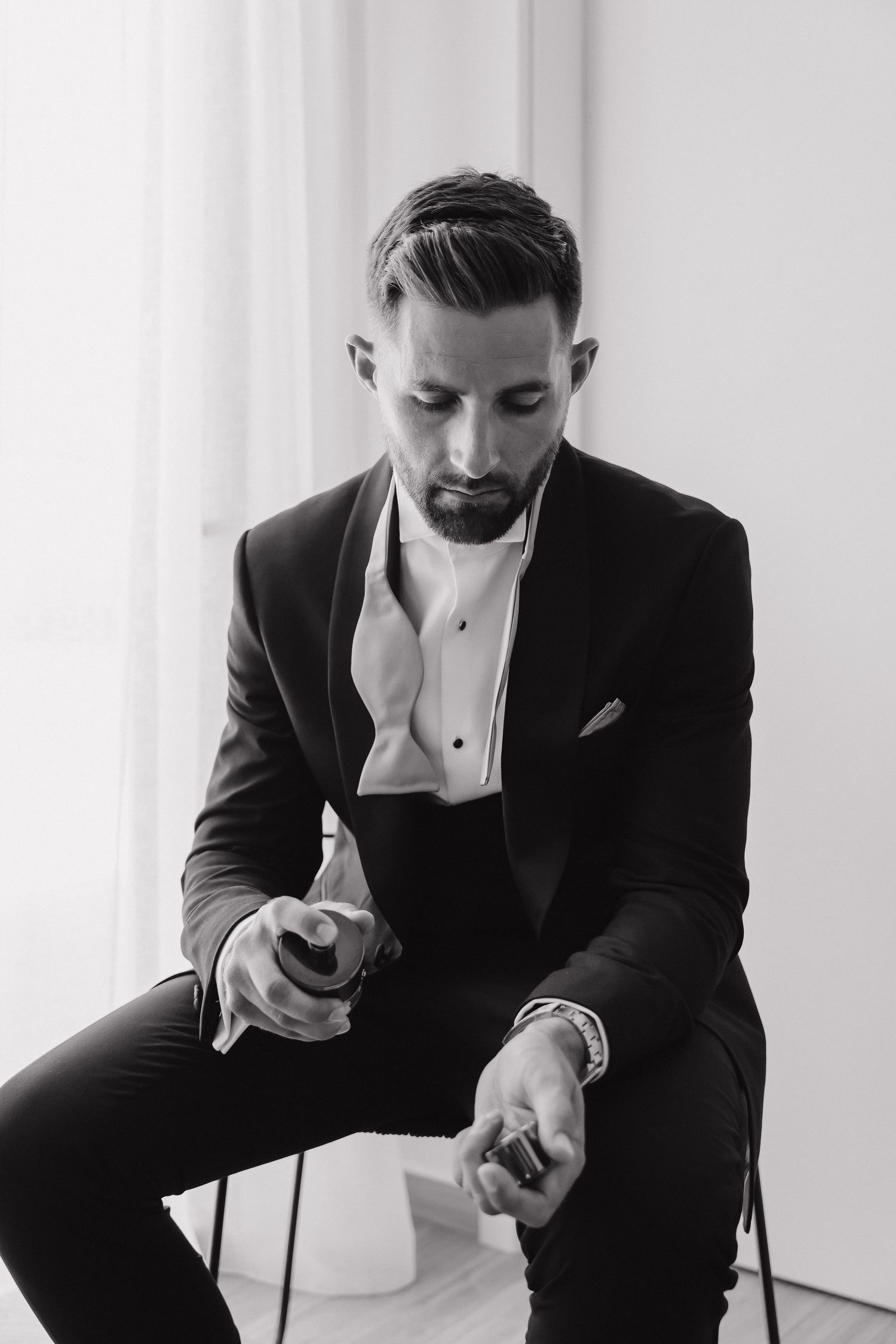 Monochrome portrait of a person in formal black tuxedo seated in minimalist setting.