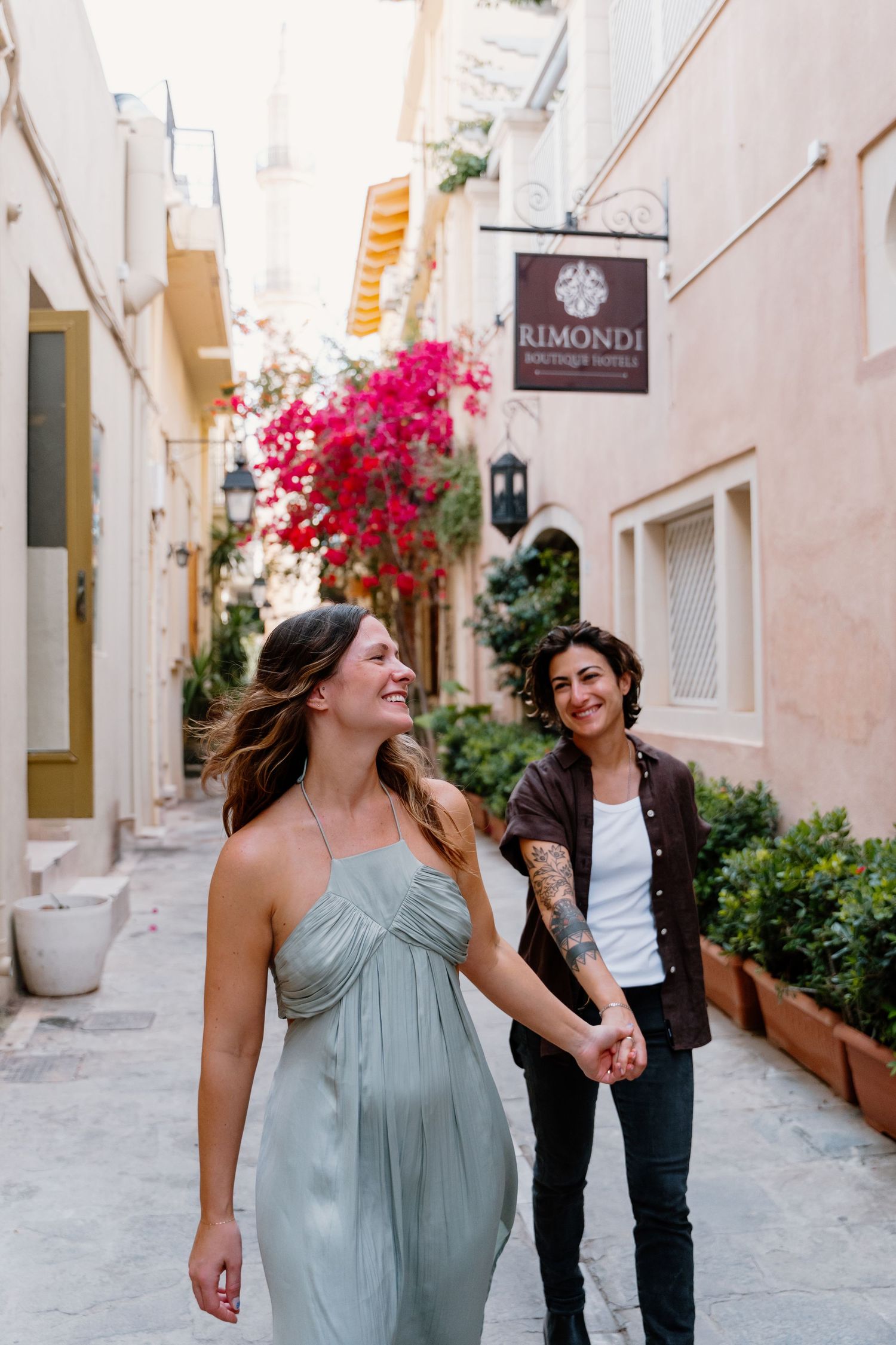 Two people laughing and walking down a charming Mediterranean alley in Rethymno, Crete with pink bougainvillea and storefronts.