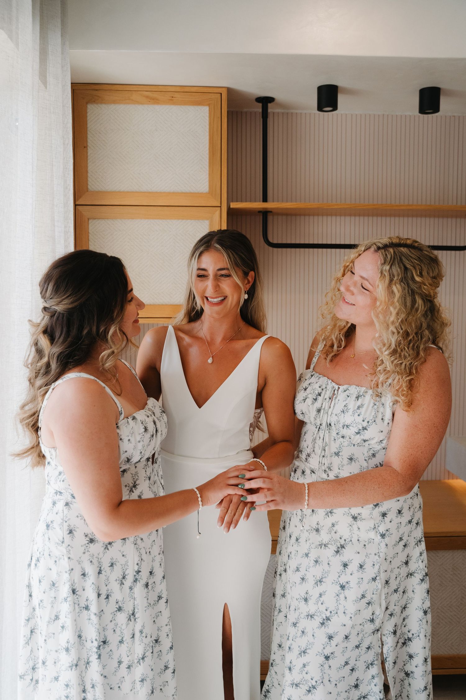 Three women in floral and white dresses share a joyful moment during wedding preparations.