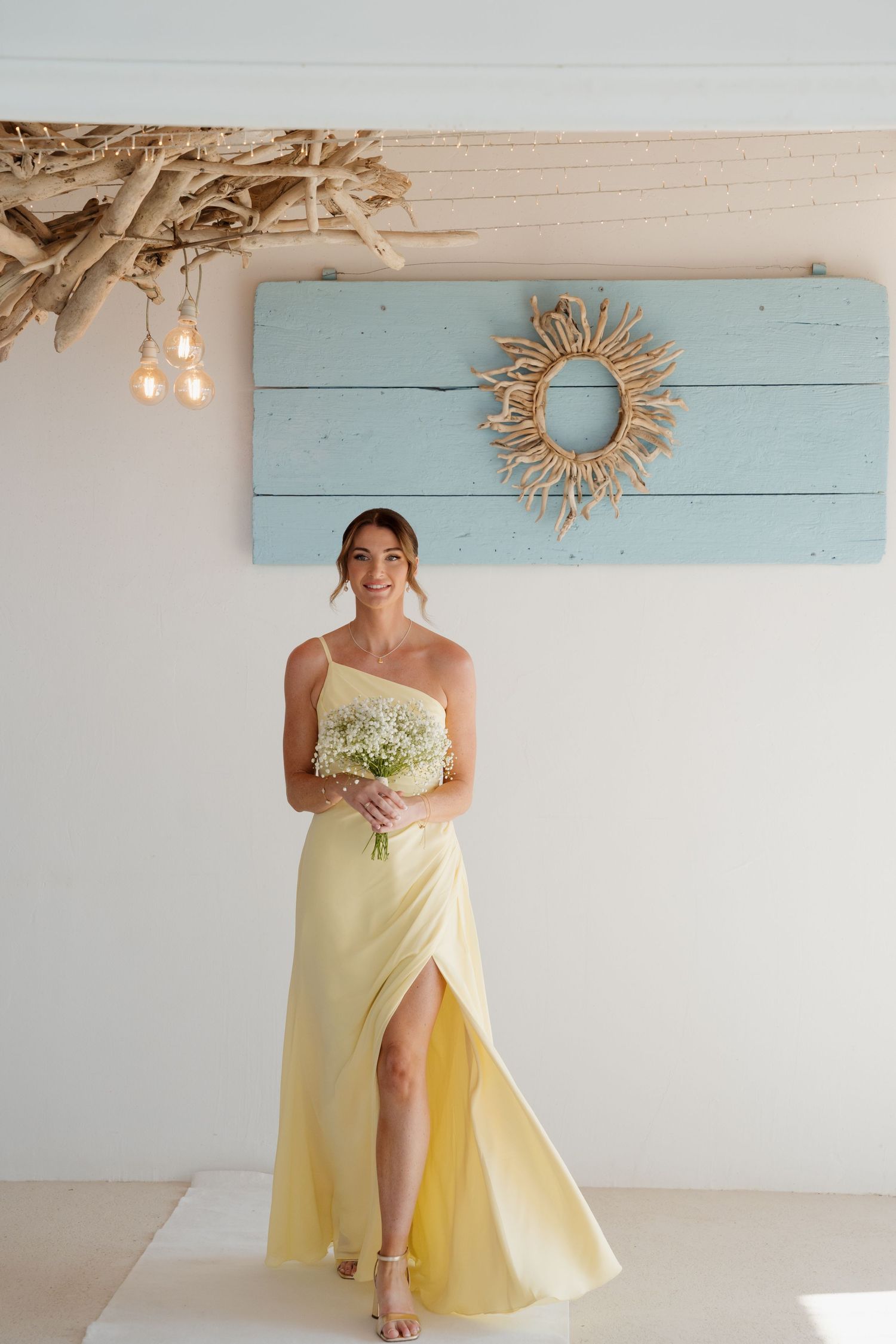 Bridesmaid in pale yellow one-shoulder dress holding baby's breath bouquet against blue wall decor.