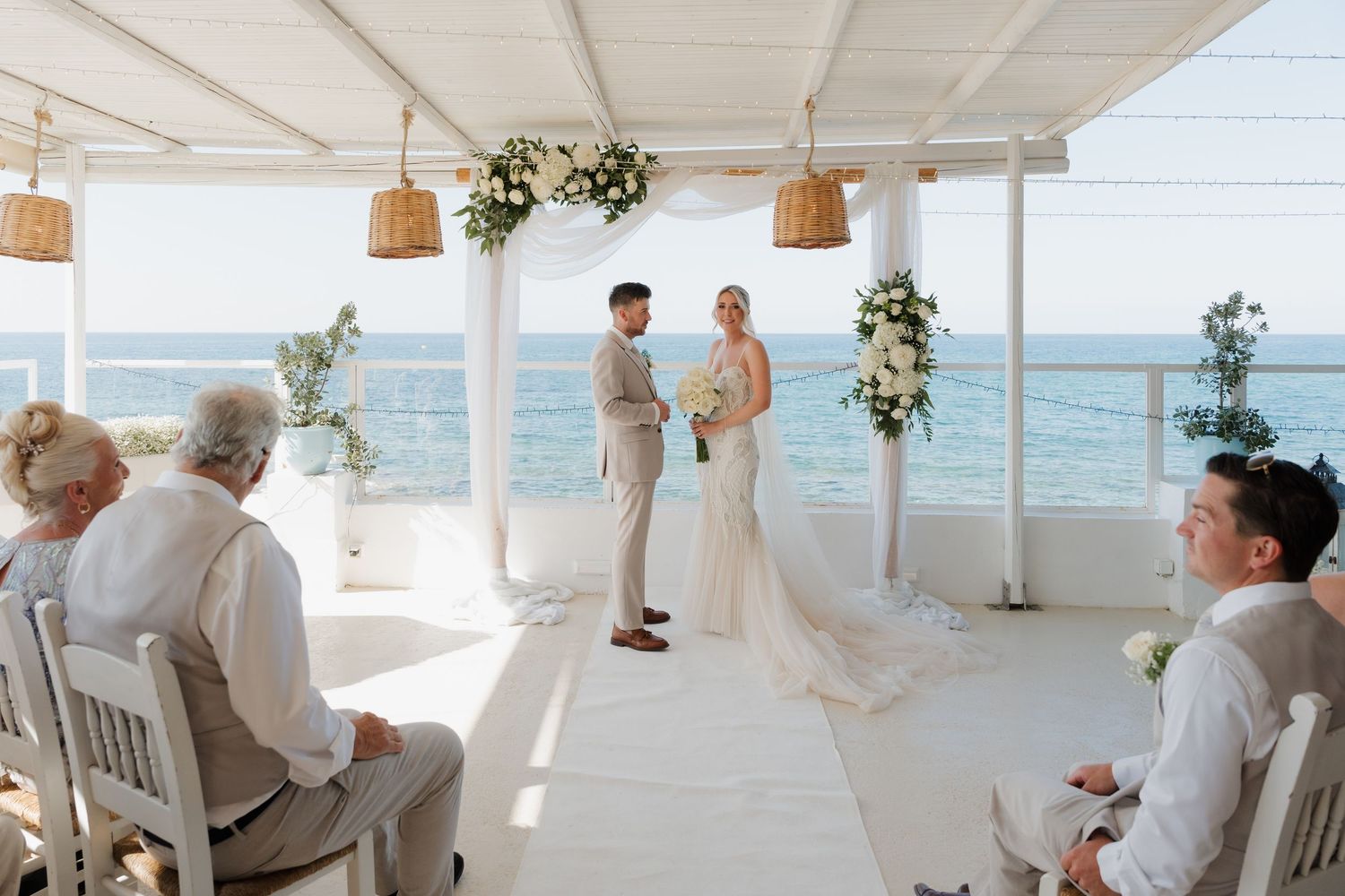 Intimate beachfront wedding ceremony with white decor and hanging basket lights.