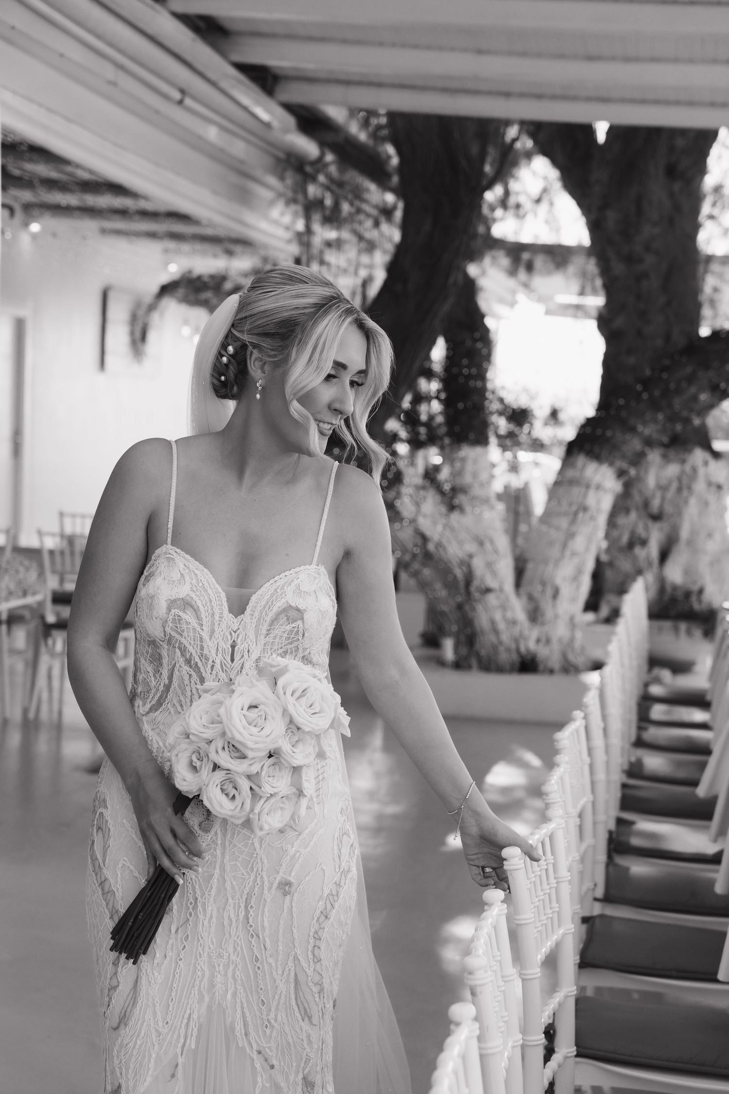 A woman in an elegant white lace wedding dress stands on a Mediterranean balcony in black and white photography.