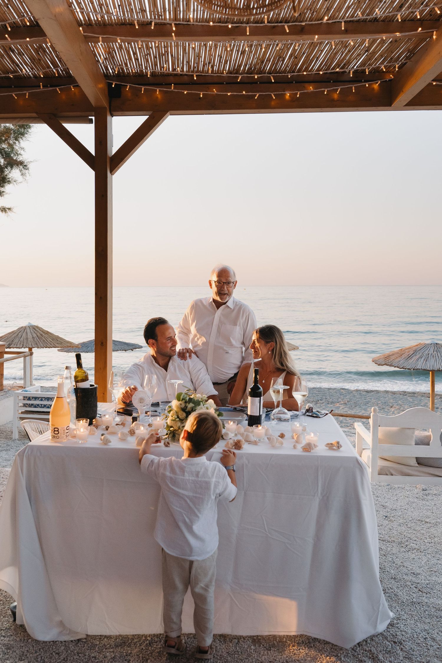 A sunset beachside reception with guests dining under a wooden pergola overlooking the ocean.