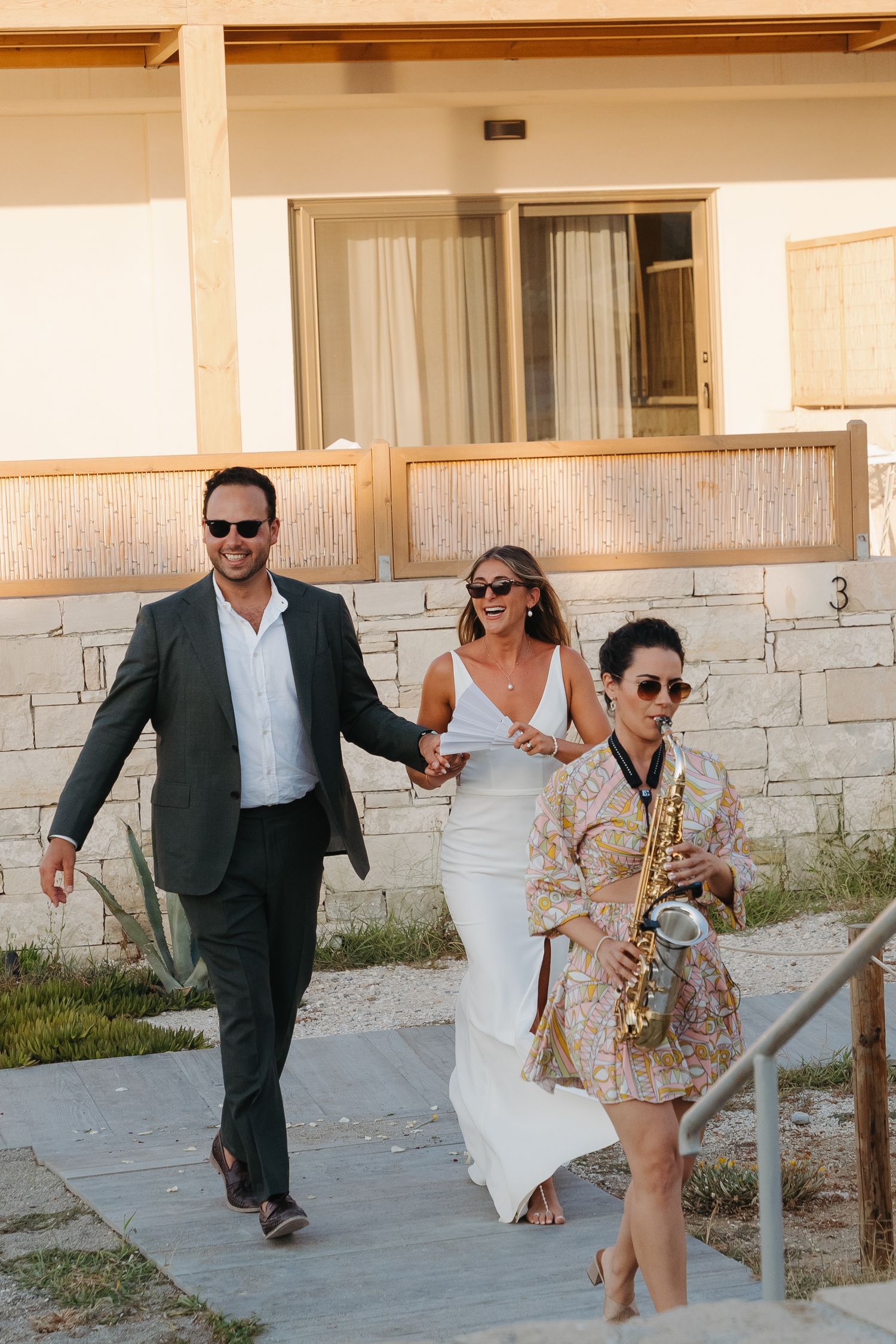Wedding party strolling down sunny pathway with saxophonist in floral dress.