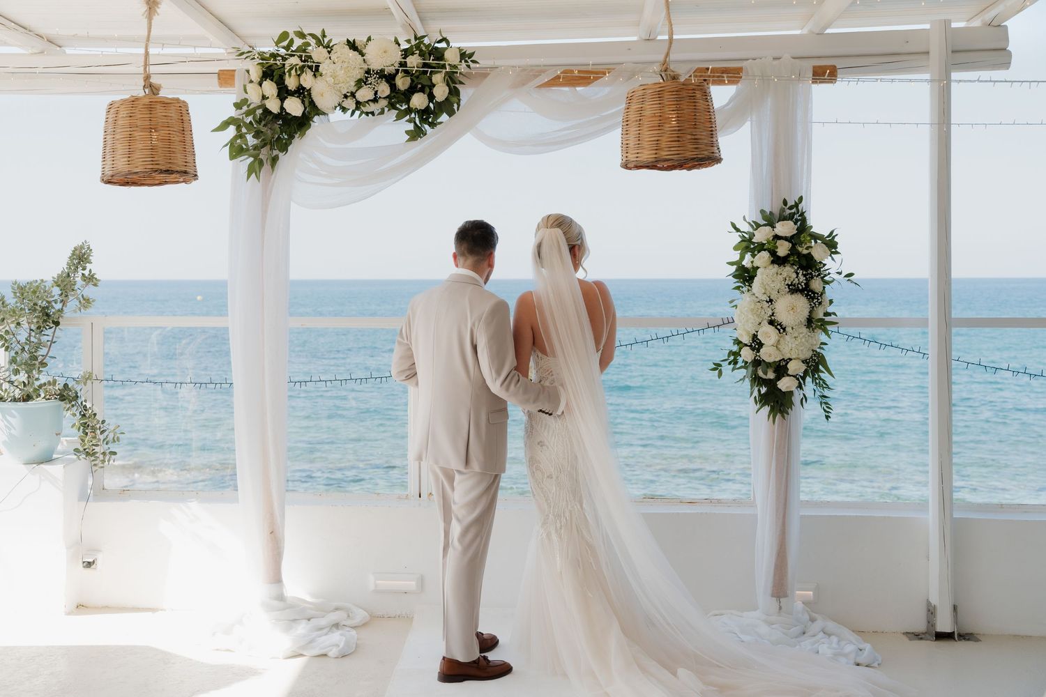 Couple stands at beachfront altar decorated with white flowers and draped fabric.