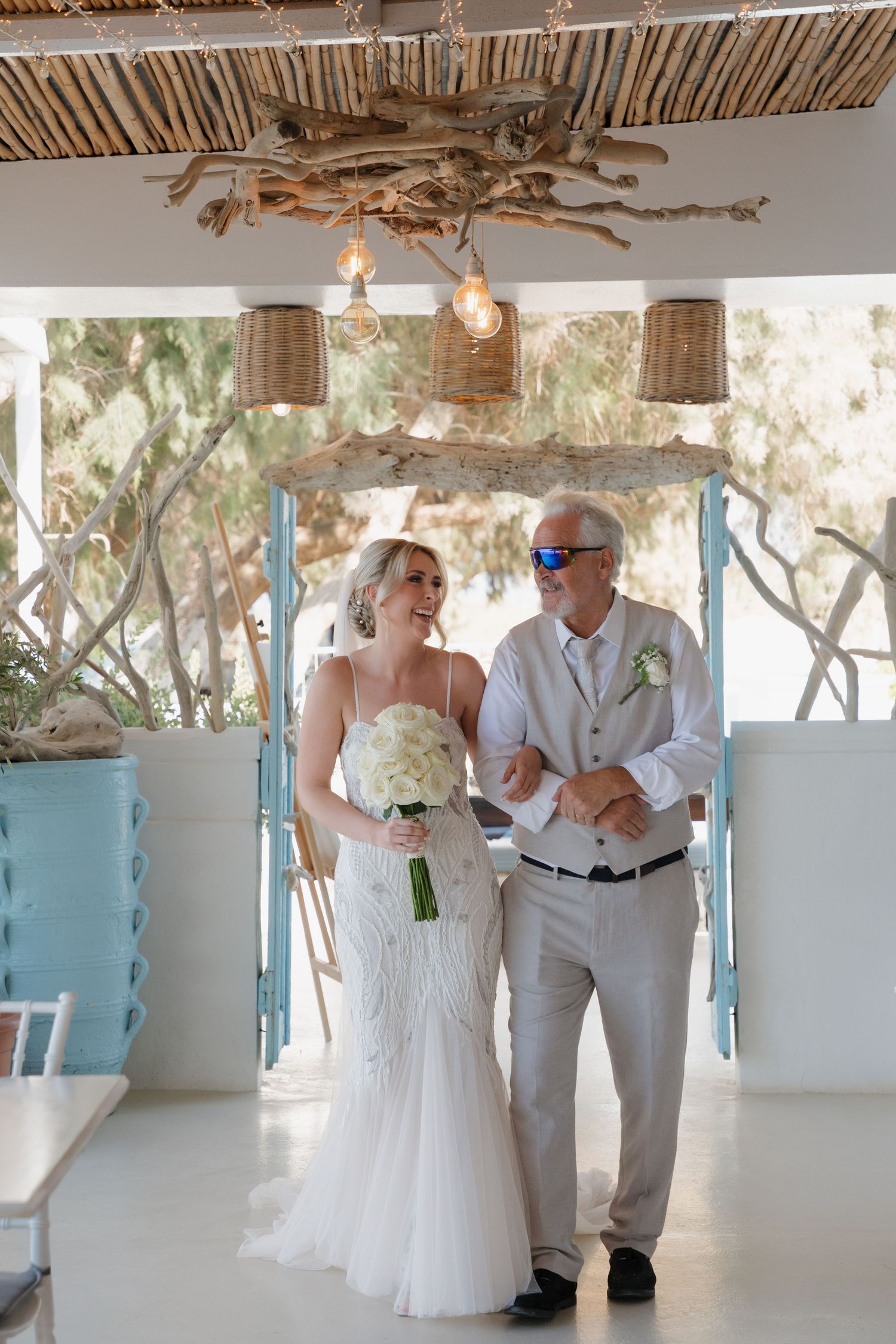 Two people in wedding attire walk under rustic wooden ceiling with woven pendant lights.