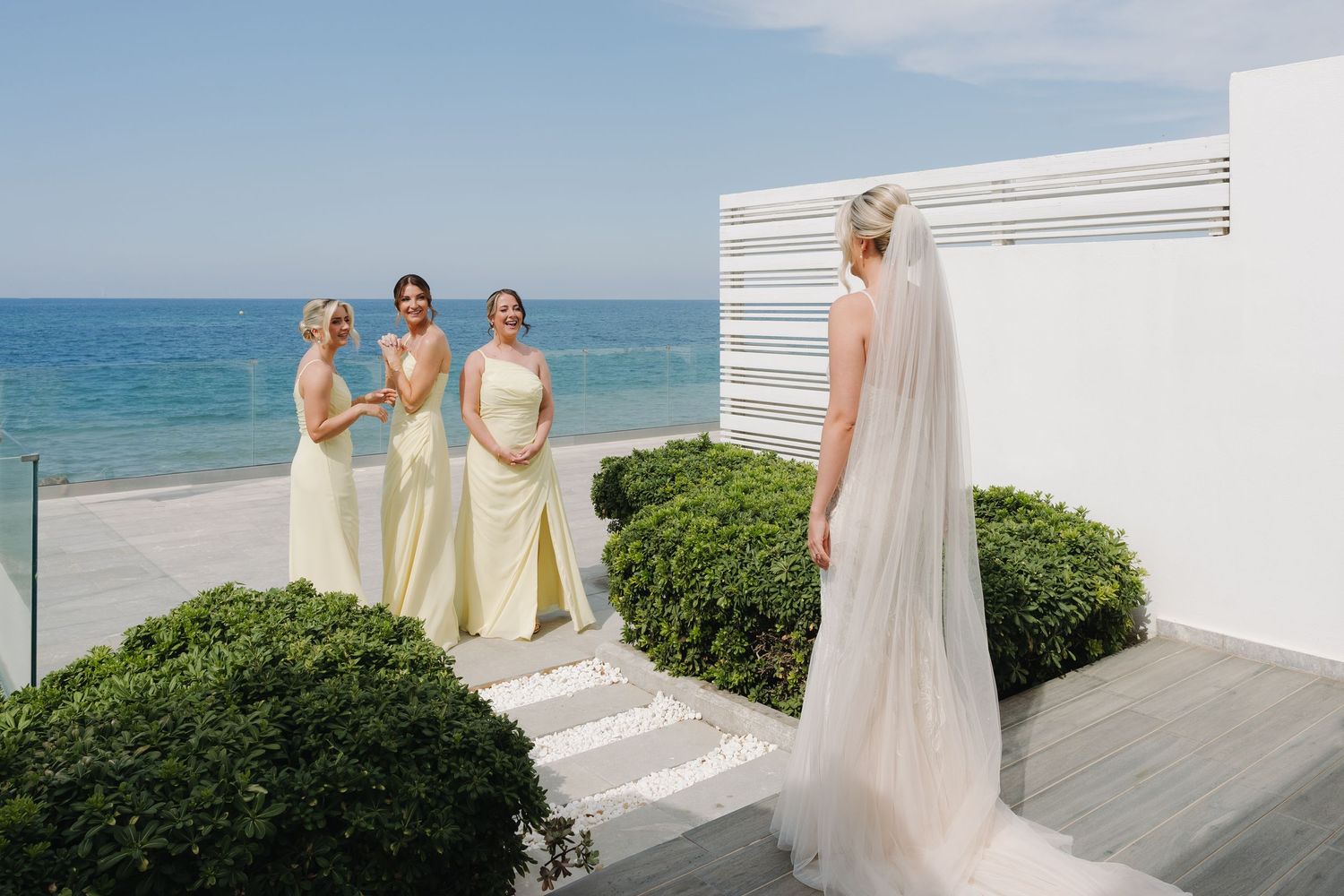 Yellow-dressed bridesmaids watching figure in flowing veil against ocean backdrop.