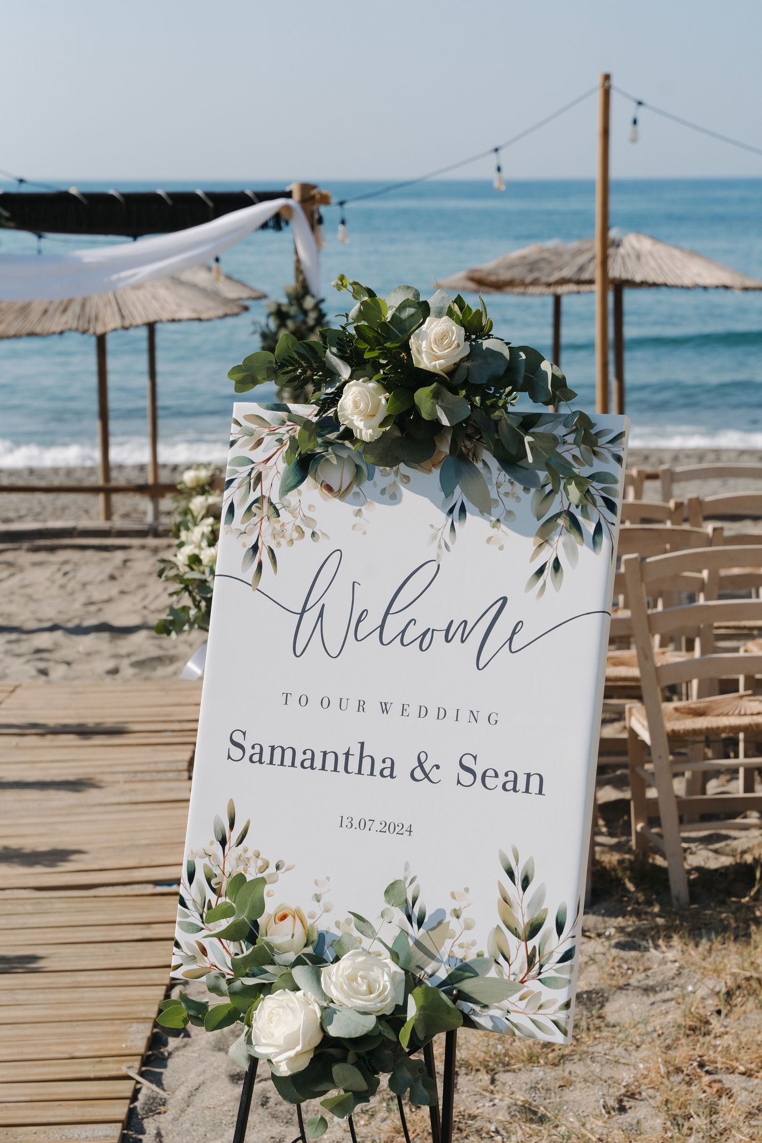 Welcome sign decorated with white roses and greenery stands on a beach boardwalk with umbrellas and ocean views.