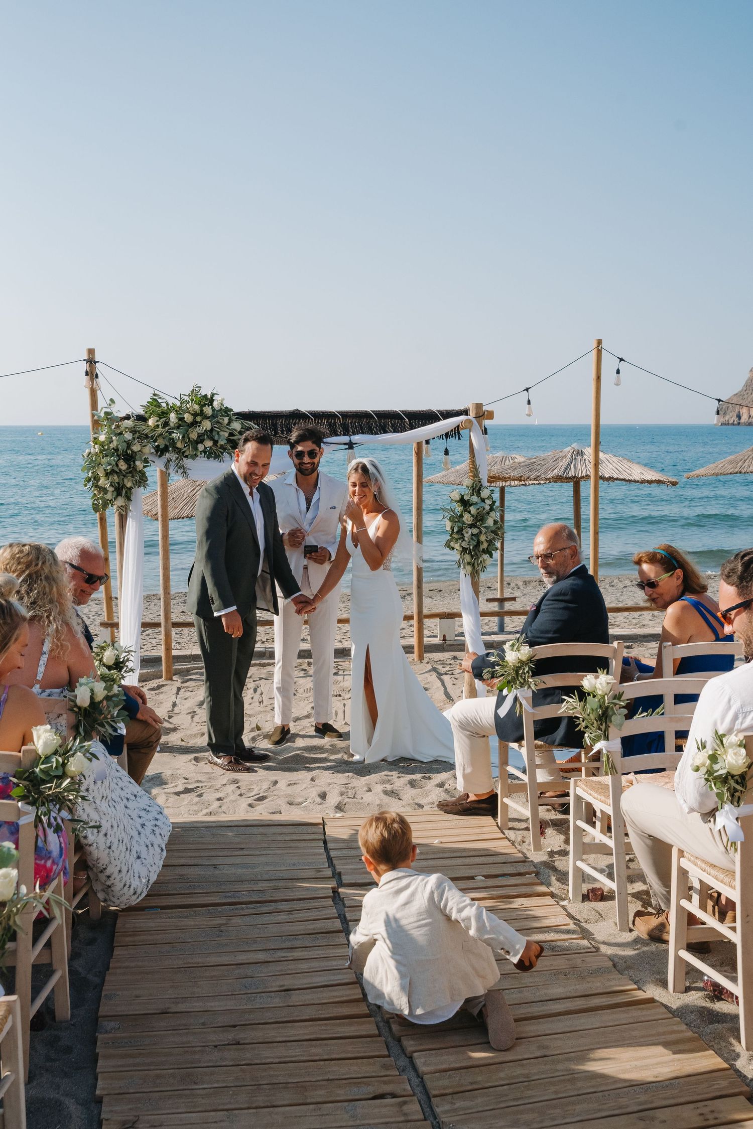 Beachside wedding ceremony taking place on wooden boardwalk with ocean backdrop and floral arch.
