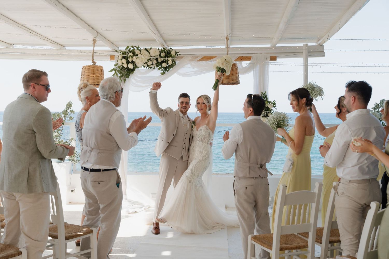 Wedding celebration with guests cheering during reception under white canopy by ocean.