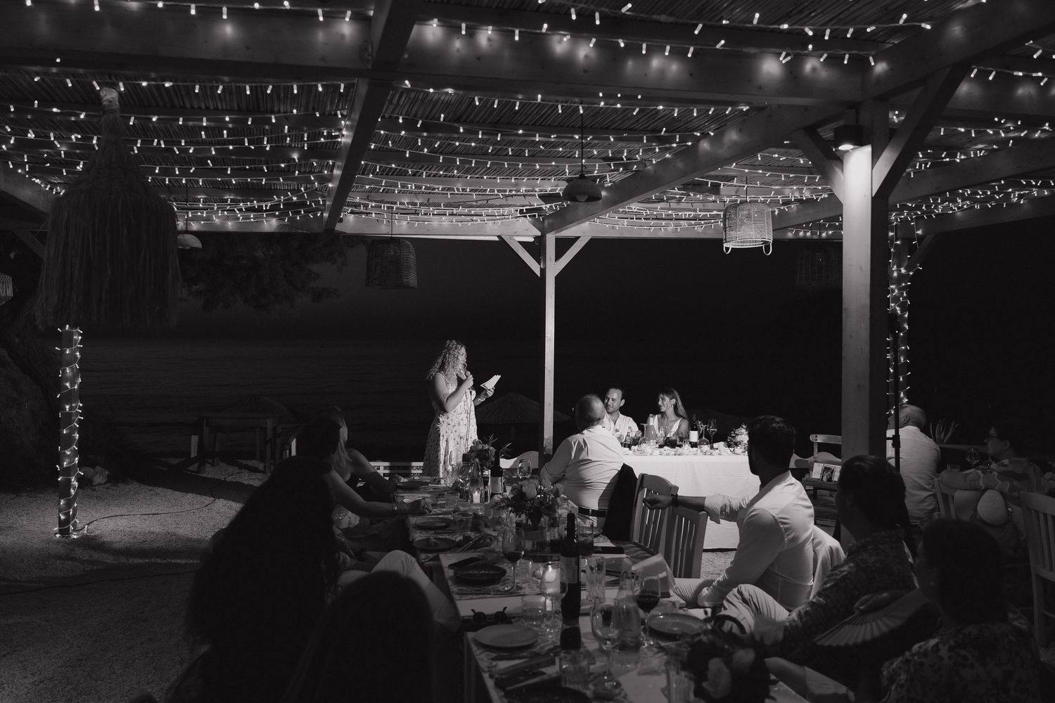 A black and white reception photo showing guests dining under twinkling lights at night.