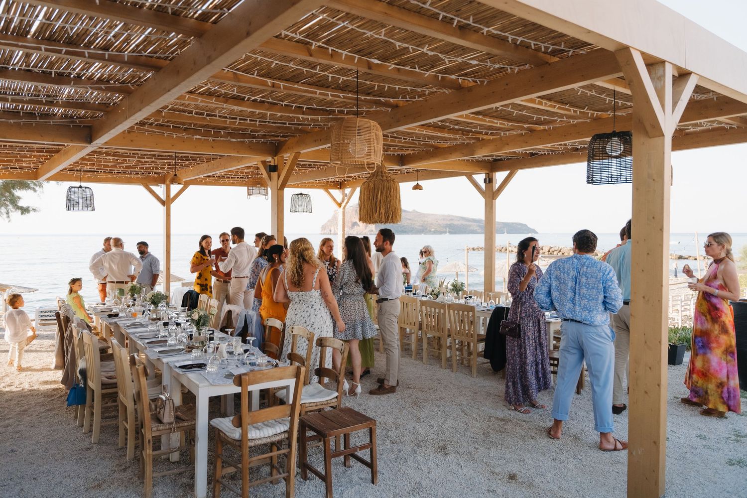 Beachside wedding reception setup with wooden pergola and rustic Mediterranean style dining tables.