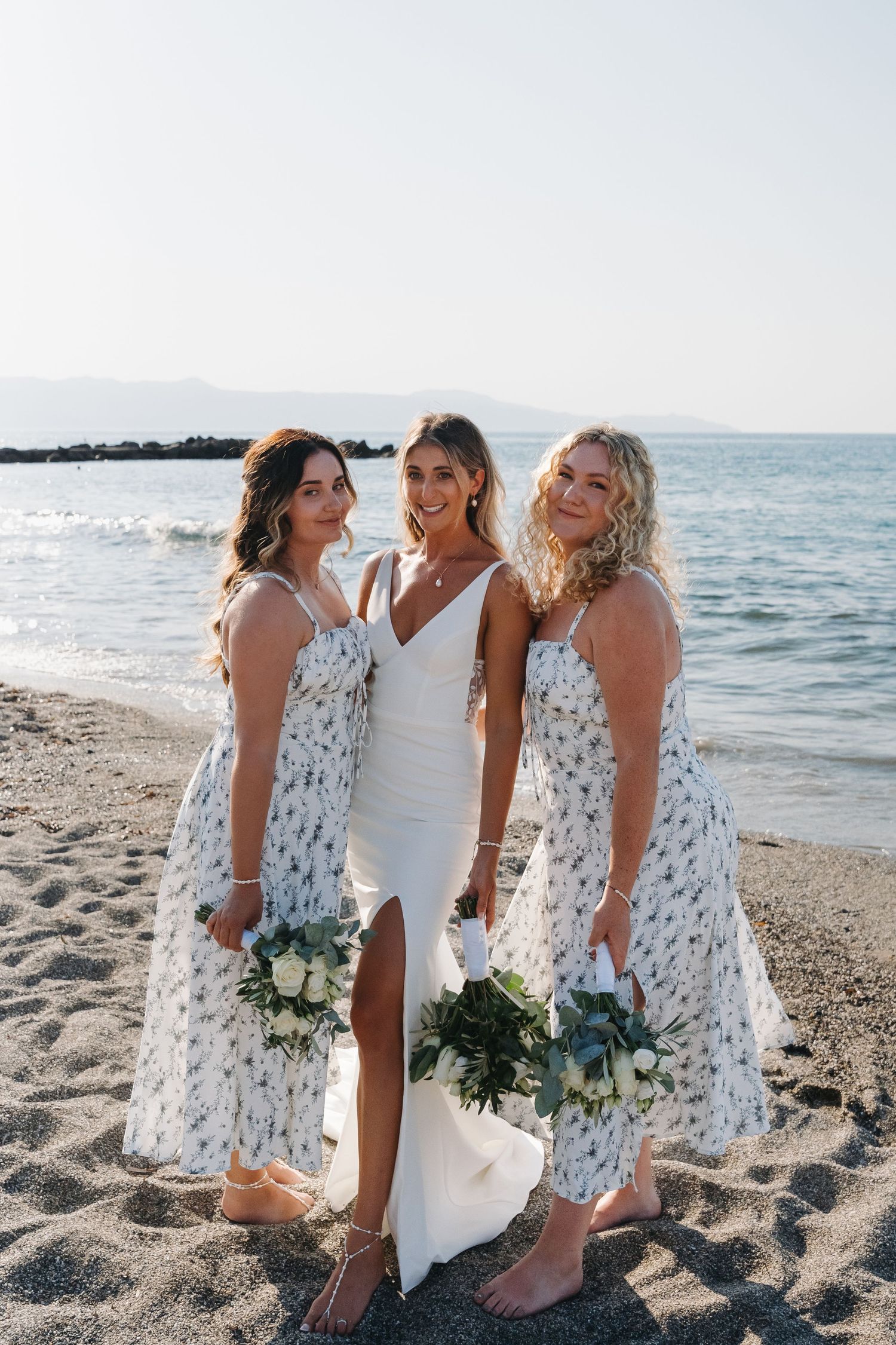 Three women in white and floral dresses pose together on a beach holding bouquets.