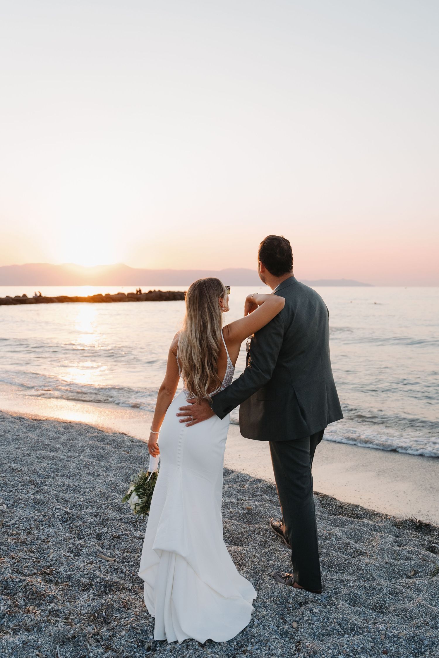 Couple watches sunset on beach in wedding attire while embracing.