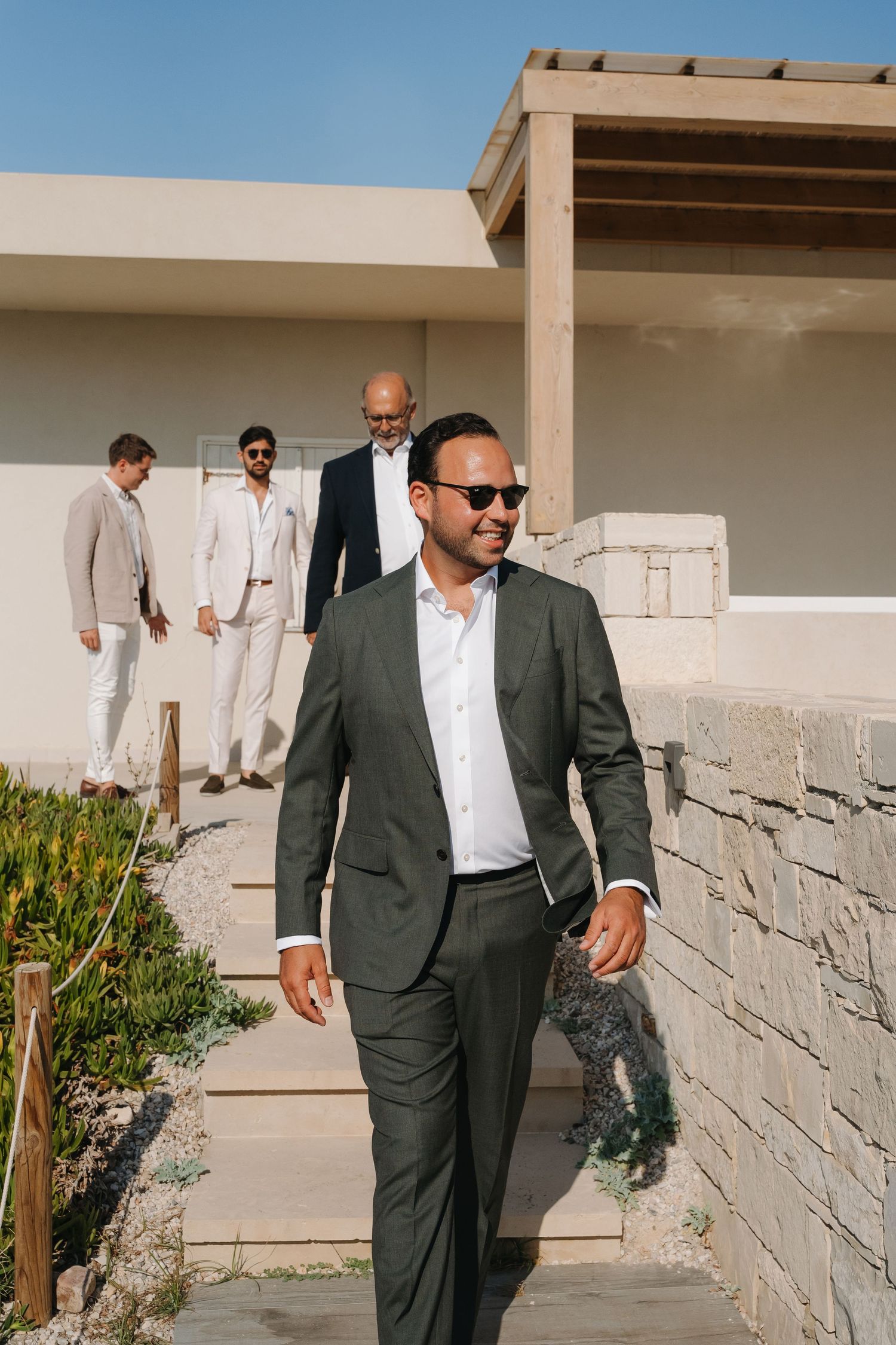 Wedding party members in stylish suits walk down outdoor steps at a sunny venue.