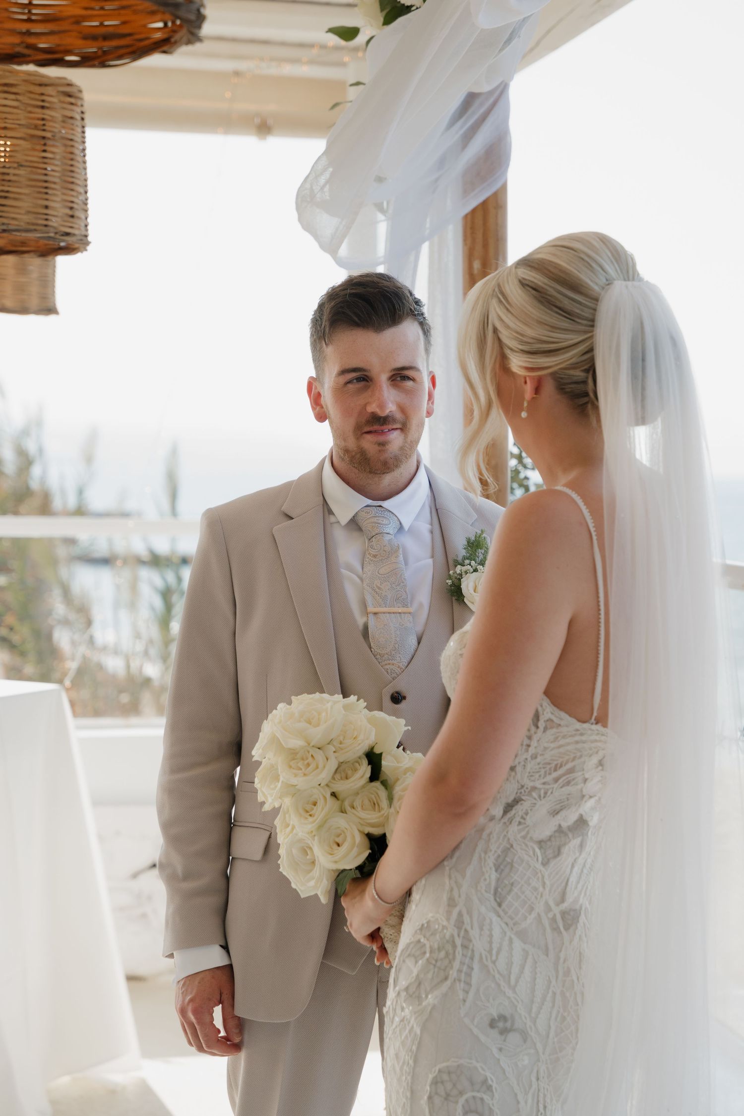 Intimate beachfront wedding ceremony with white decor and woven pendant lights.