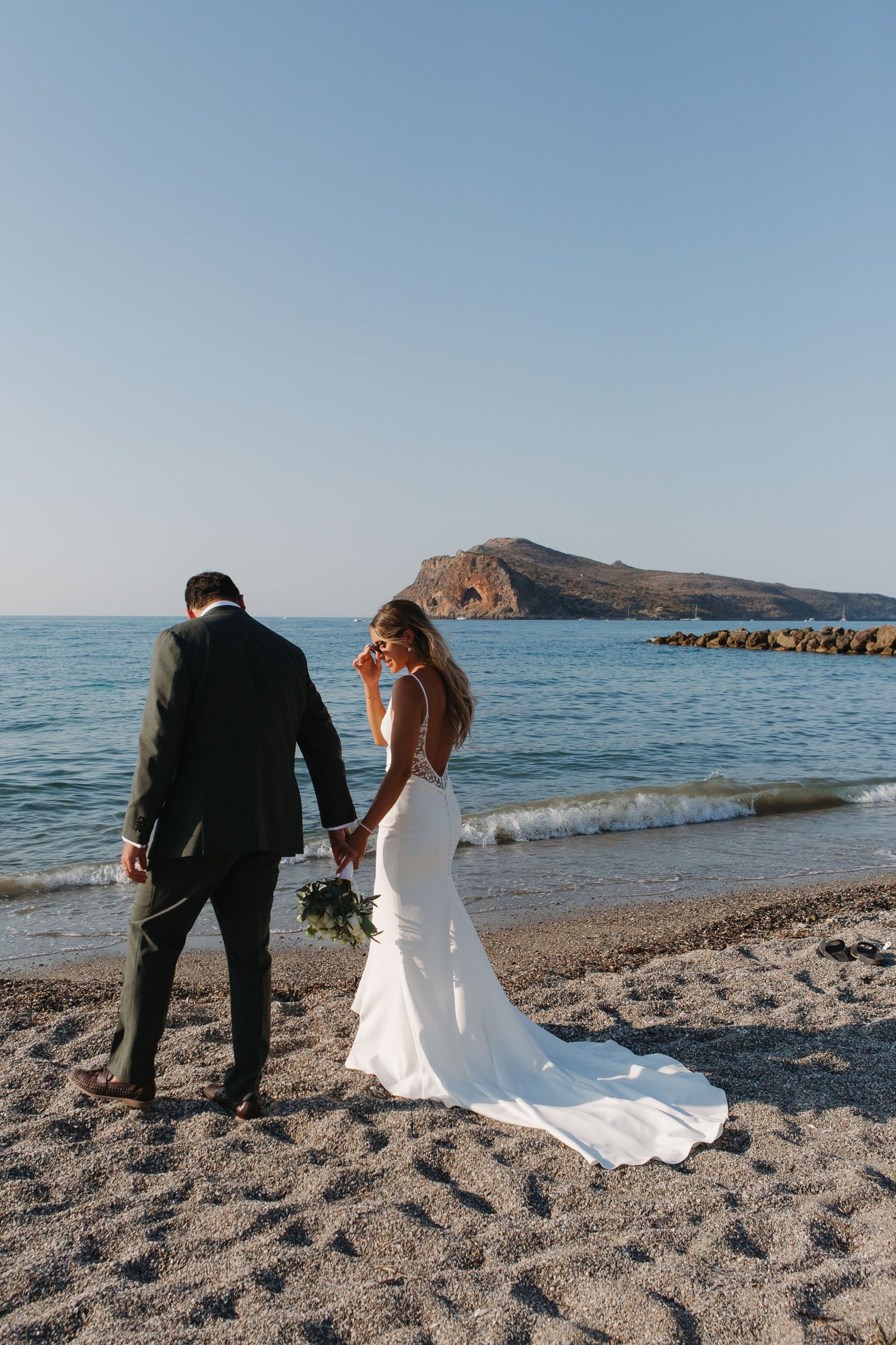 Couple walks along a scenic Mediterranean beach shoreline during their wedding photo session.