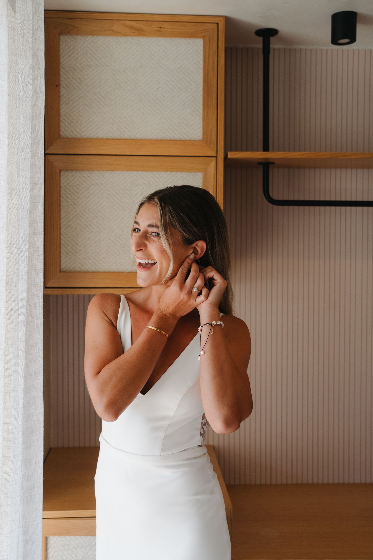 A woman in a white dress laughs joyfully while getting ready in a minimalist room with wooden cabinets.