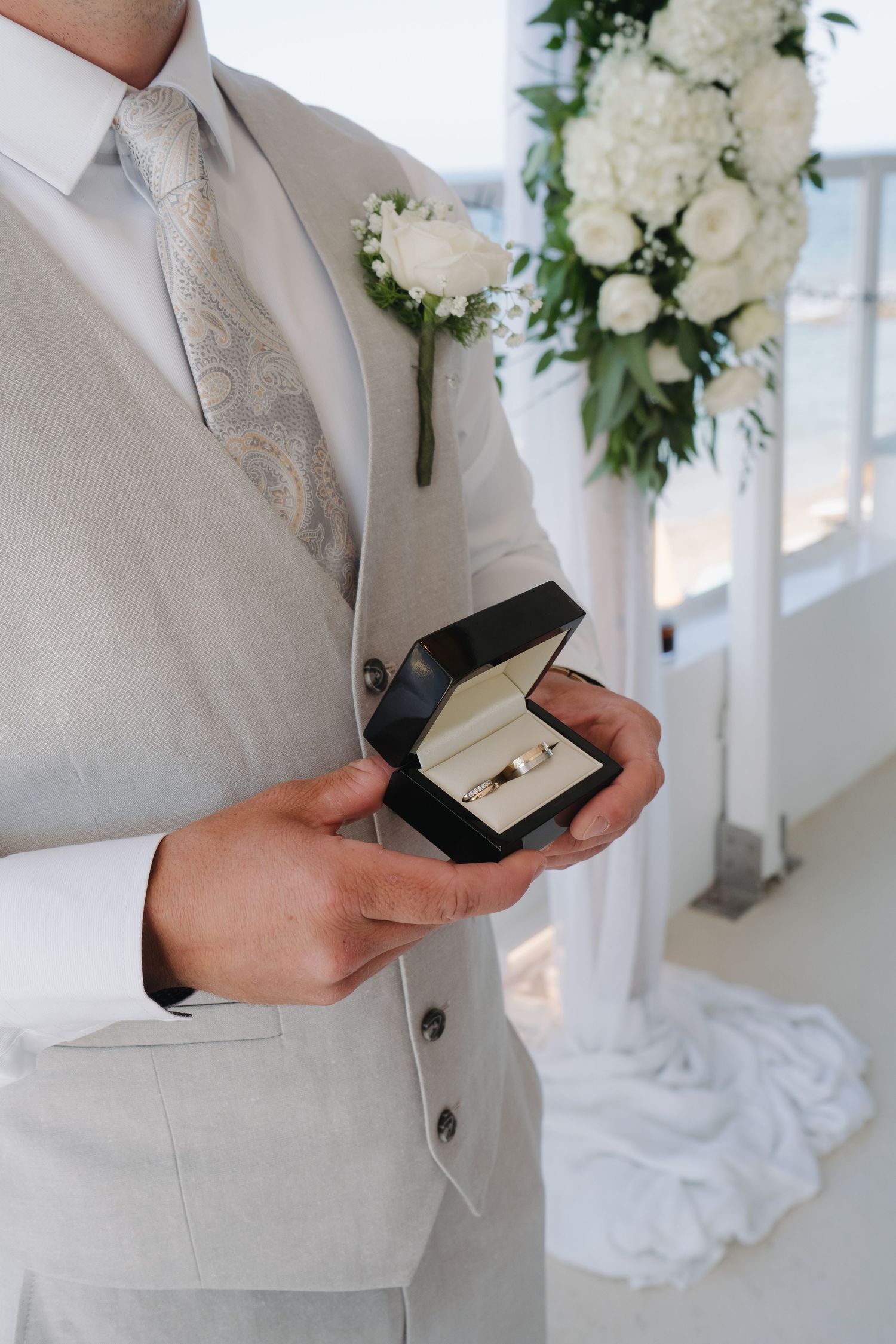 A groom in a beige suit holds an open ring box with a white floral boutonniere against an elegant wedding backdrop.