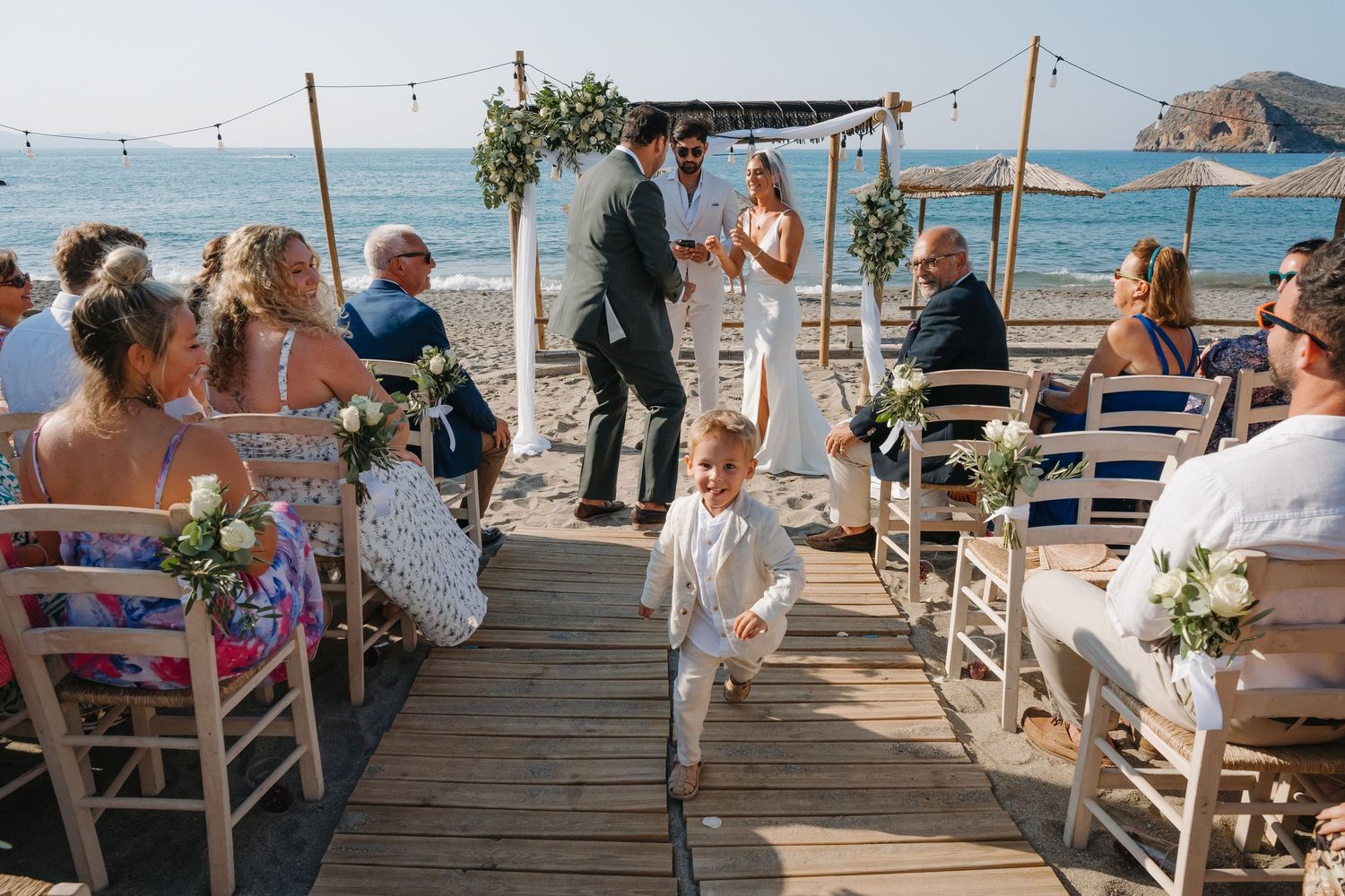 Beach wedding ceremony setup with wooden chairs and floral arch overlooking the ocean.