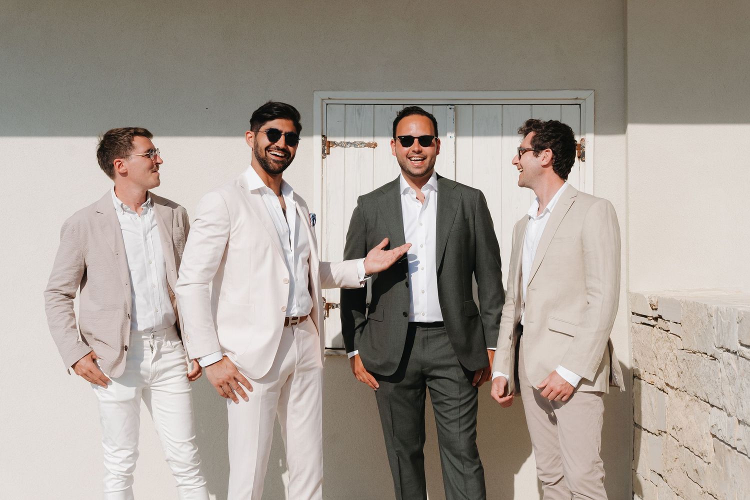 A group of groomsmen in white suits and one in gray share a candid laughing moment together at a wedding venue.