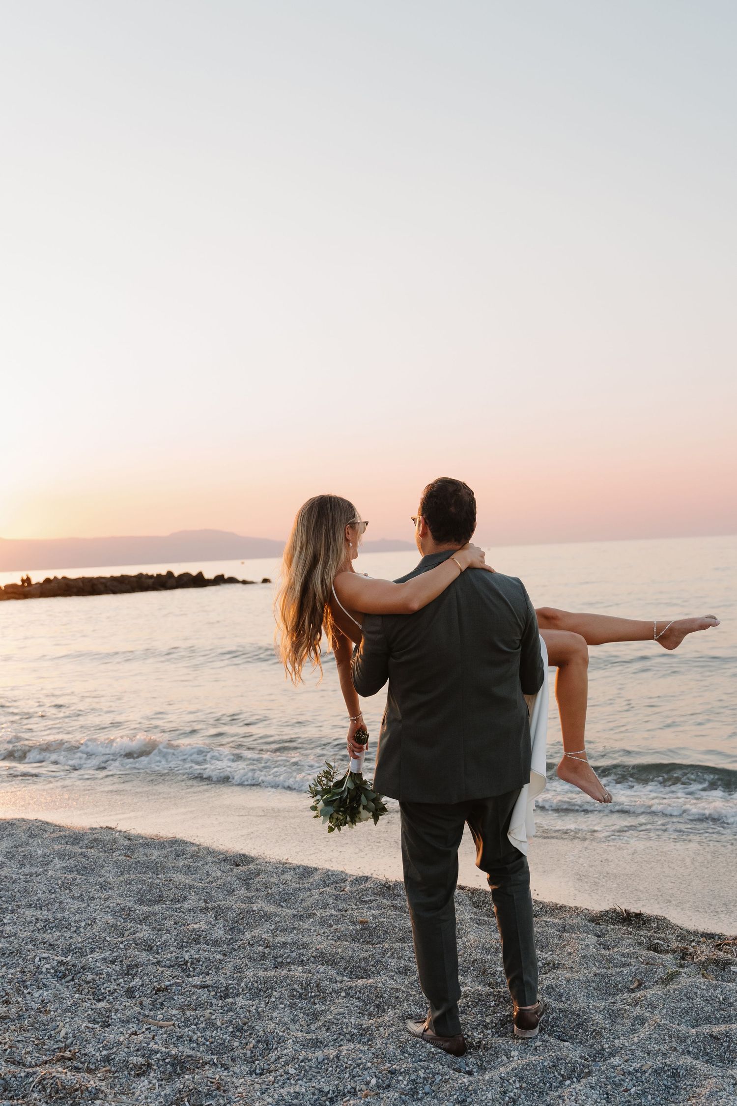 A romantic couple enjoys sunset on a Mediterranean beach with a bouquet.