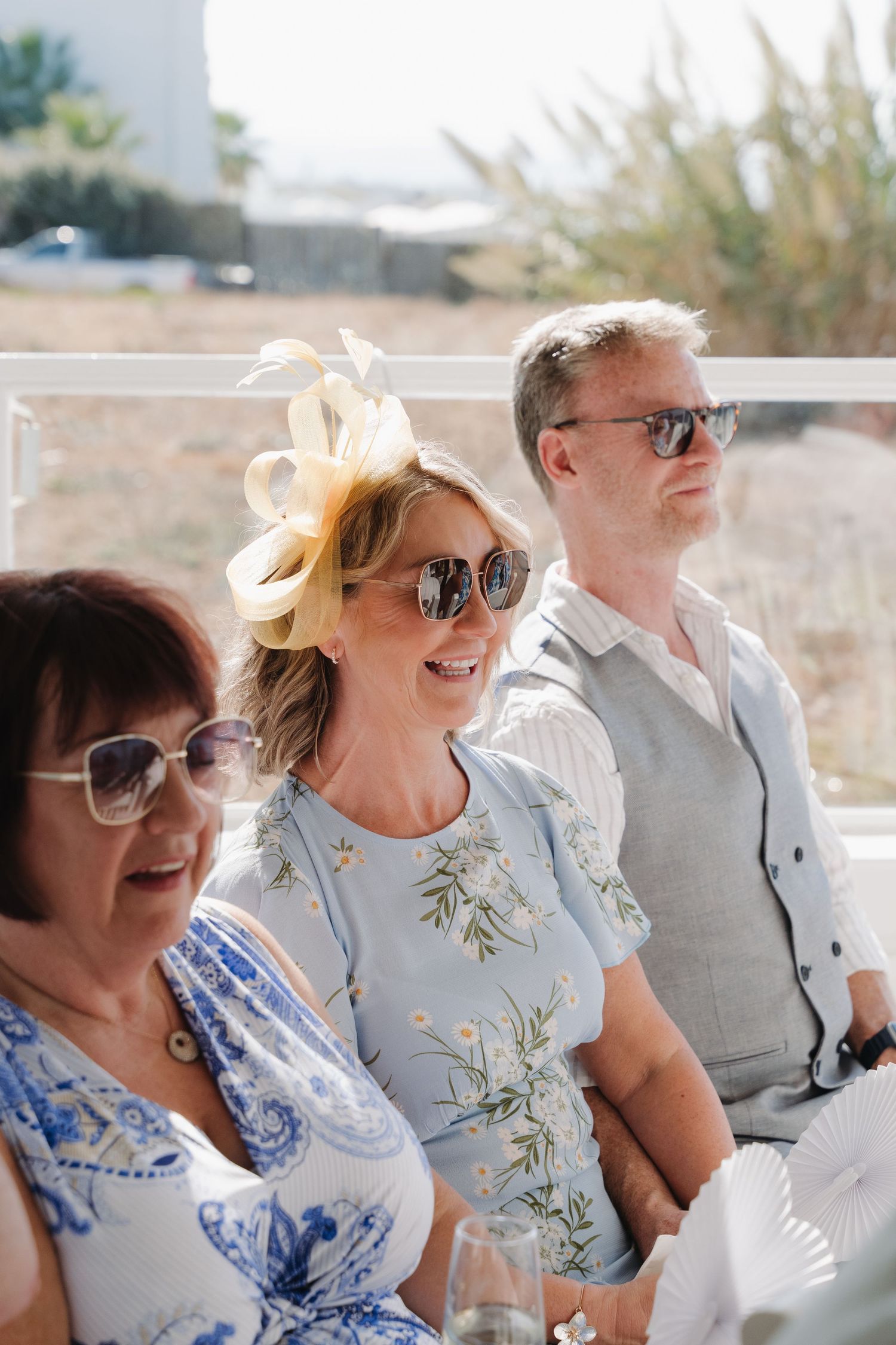 Wedding guests wearing sunglasses and summer attire smile together at an outdoor celebration.