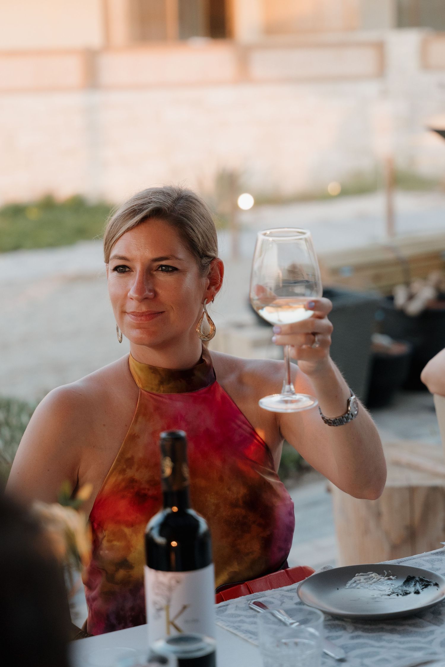 Wedding guest in a red halter dress raises a glass of wine during an outdoor reception.