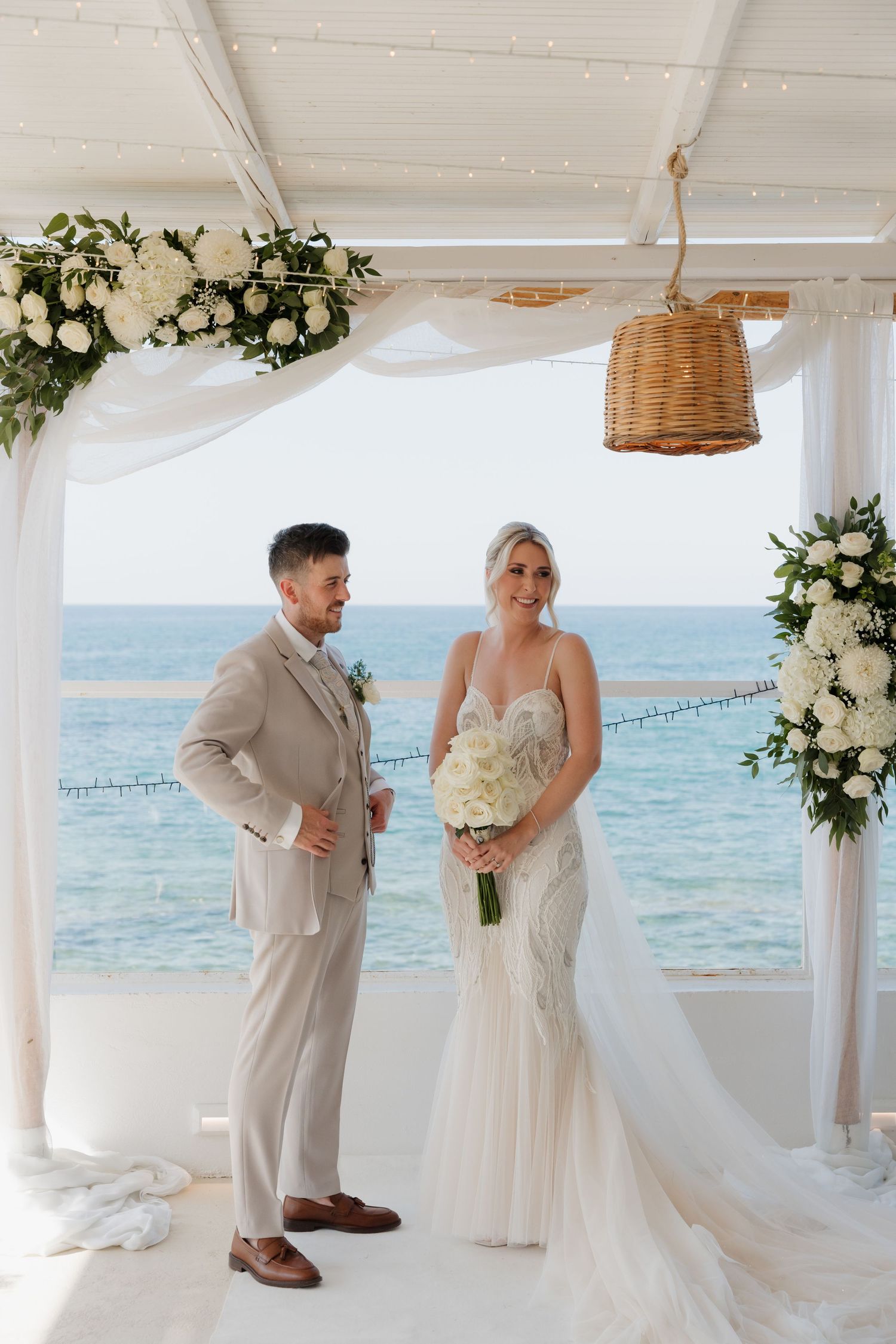 Newlyweds pose under a white pergola decorated with flowers overlooking the ocean.