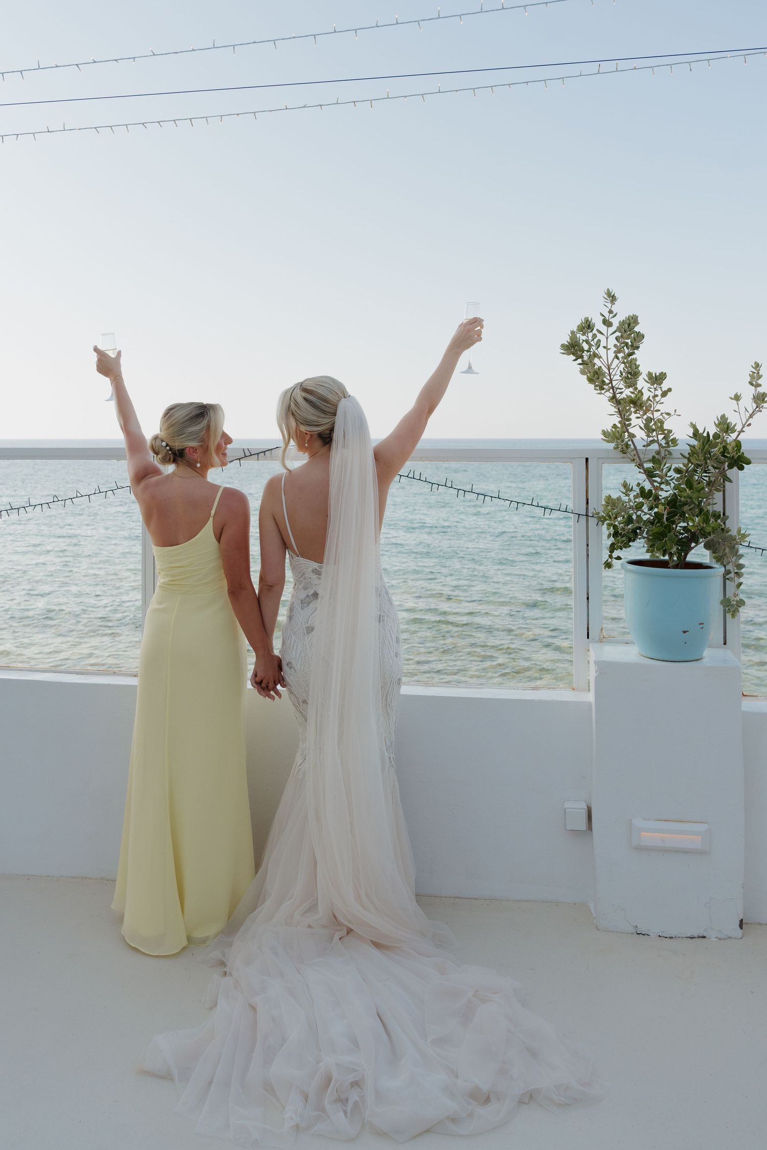 Two people in yellow and white dresses pose with arms raised on a white terrace overlooking the ocean.
