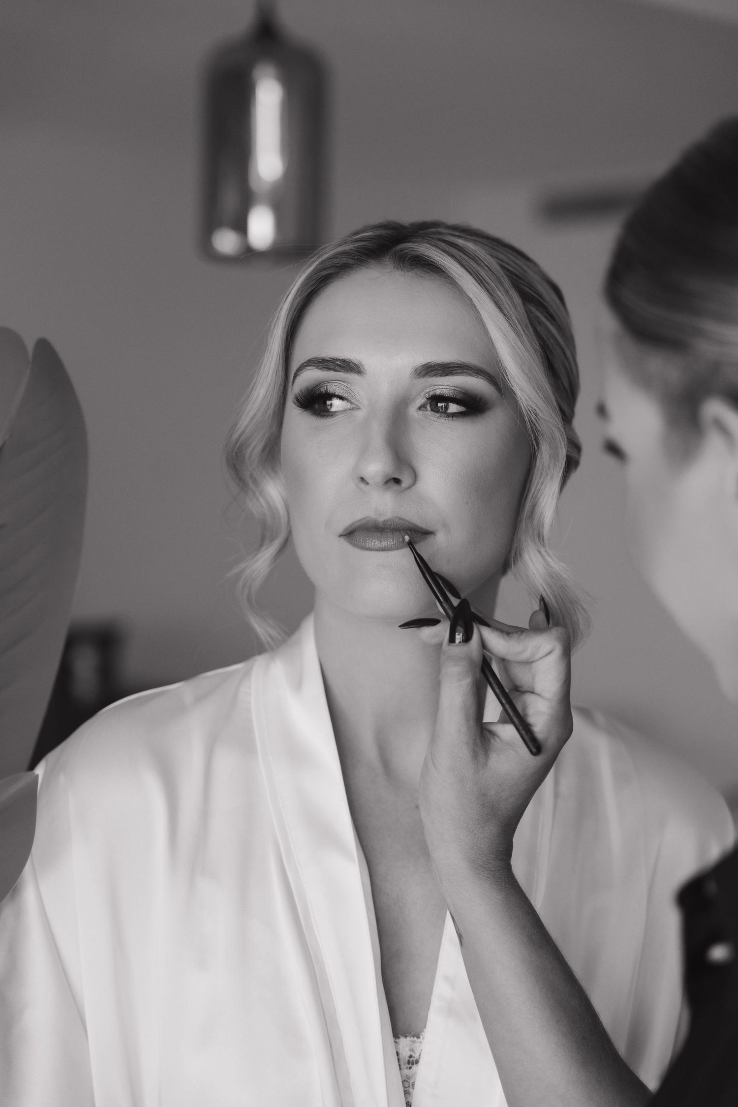 Professional makeup artist applying lip color to a bride in a white robe during wedding preparations.