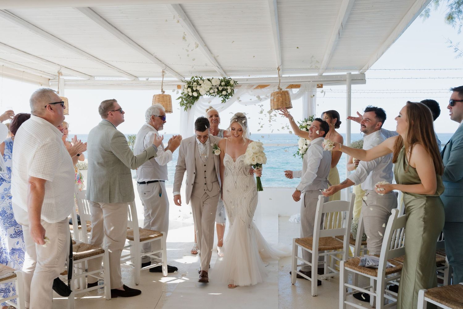 Wedding guests celebrate during a white-themed reception under a covered outdoor venue by the ocean.