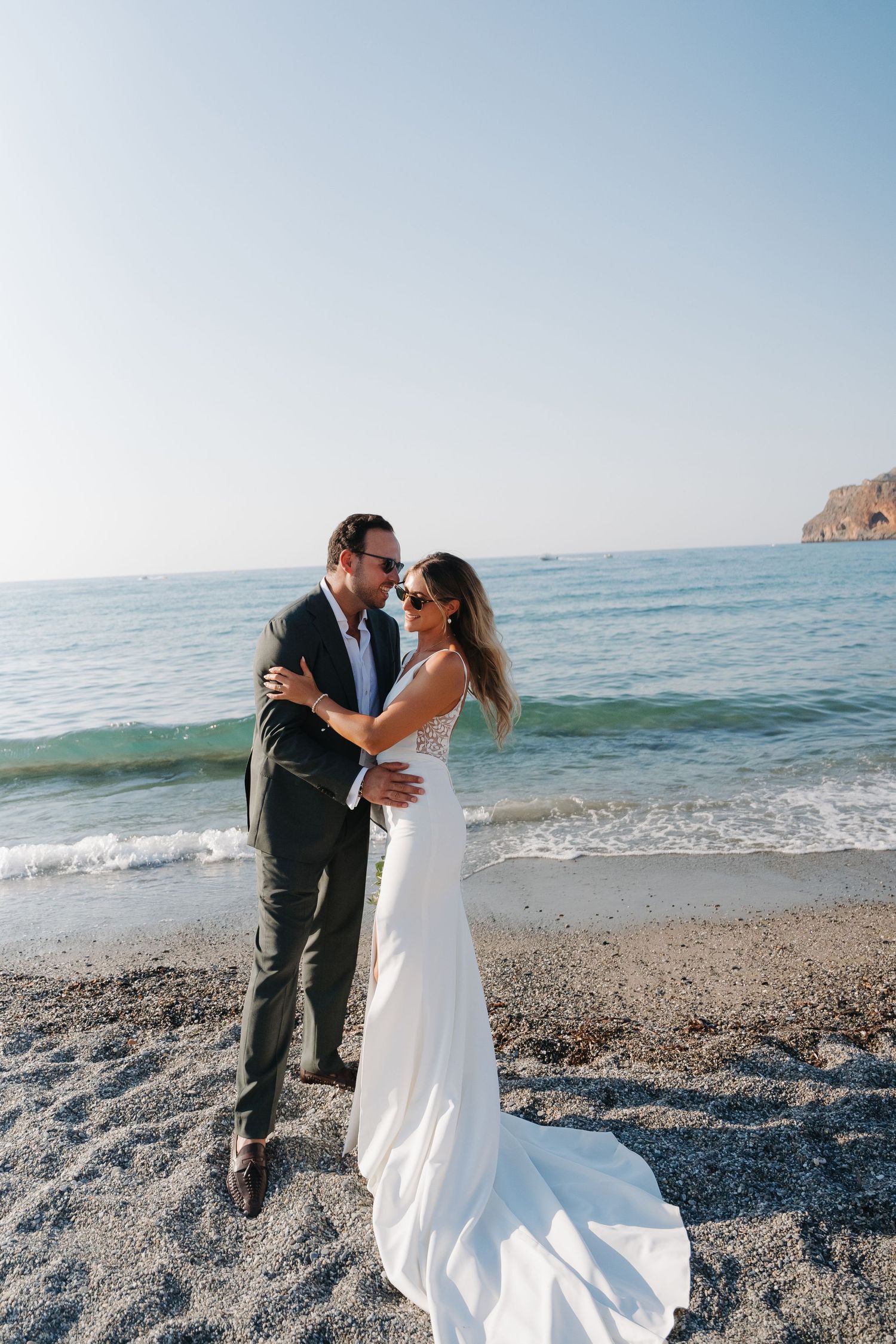 Newlywed couple embracing on beach with waves and cliffs in background.