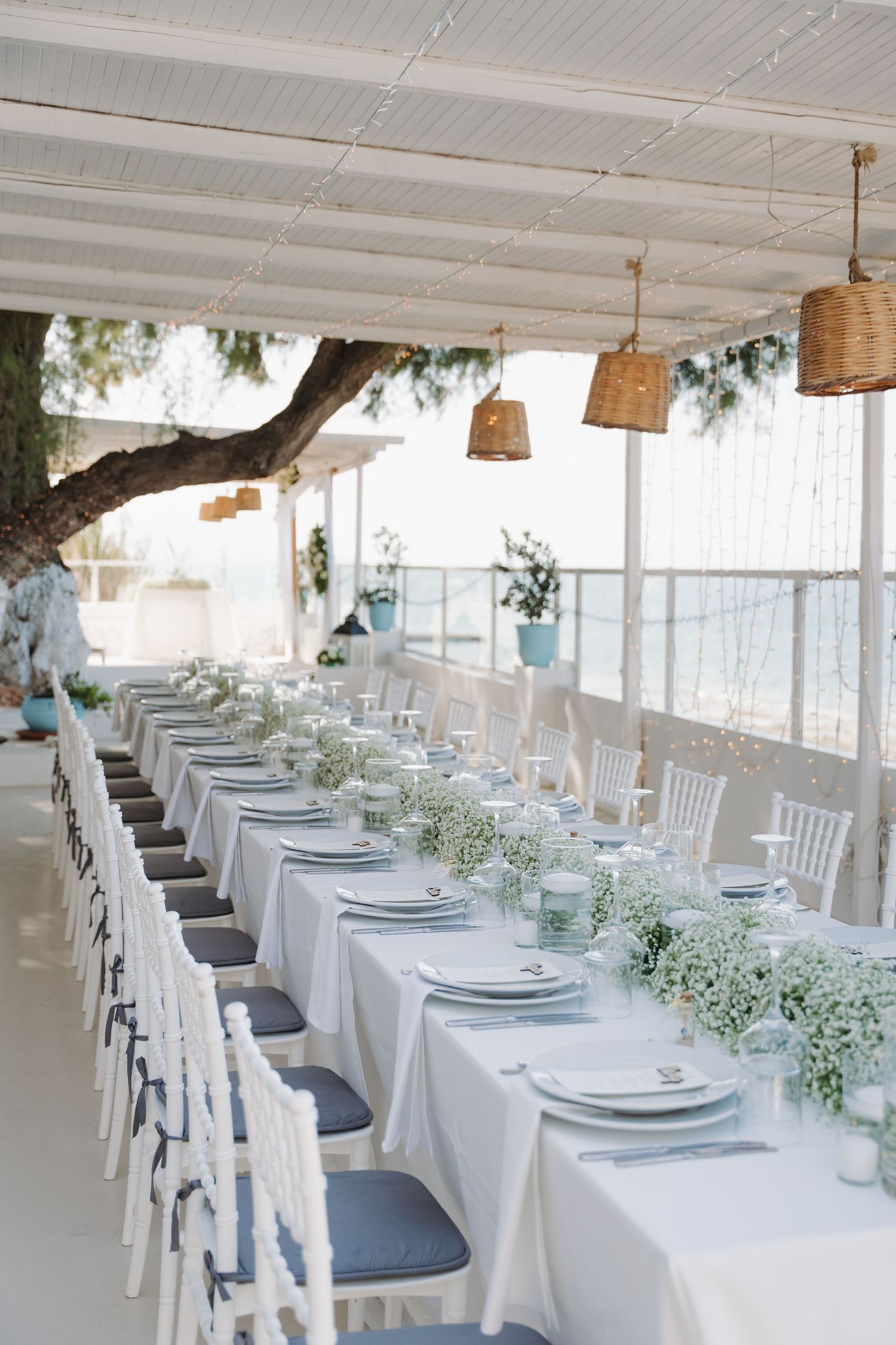 Long white wedding table decorated with floral arrangements and hanging rattan lamps near the ocean.