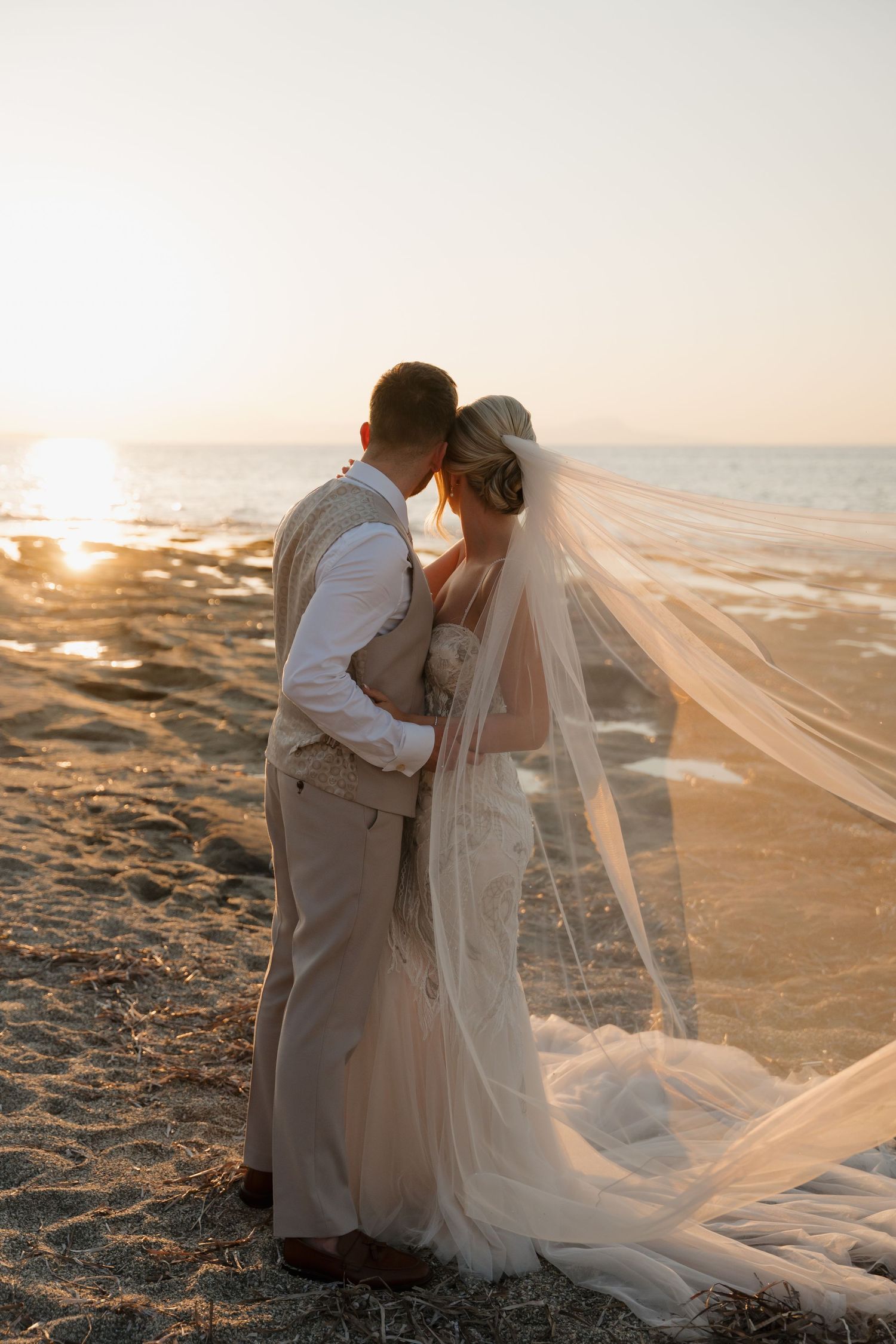 Sunset beach wedding portrait with flowing veil caught in ocean breeze.