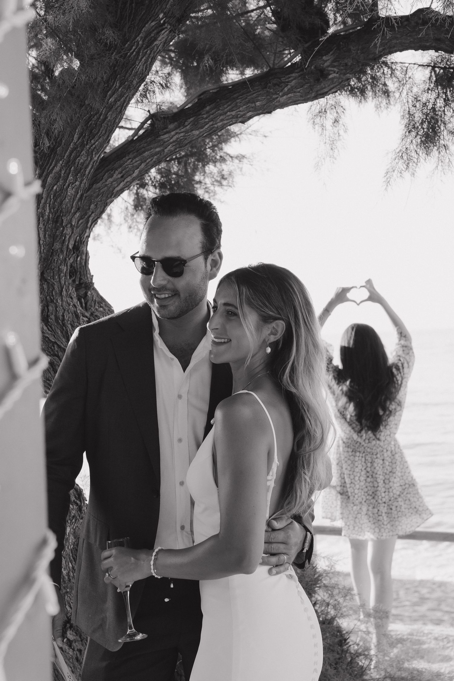 A romantic black and white beachside moment with a couple embracing under a tree at sunset.