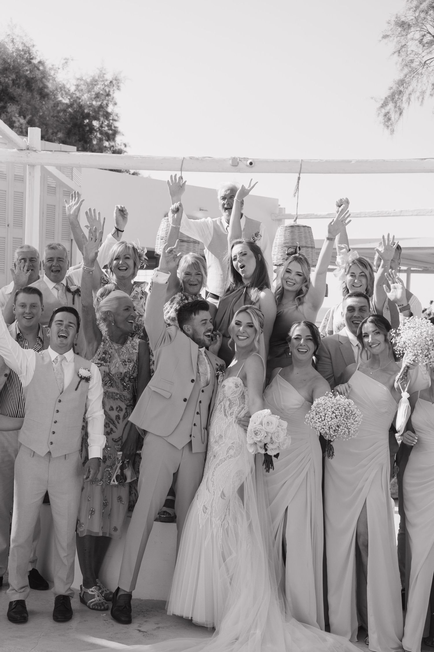 Large wedding group poses with raised arms for joyful celebration photo.