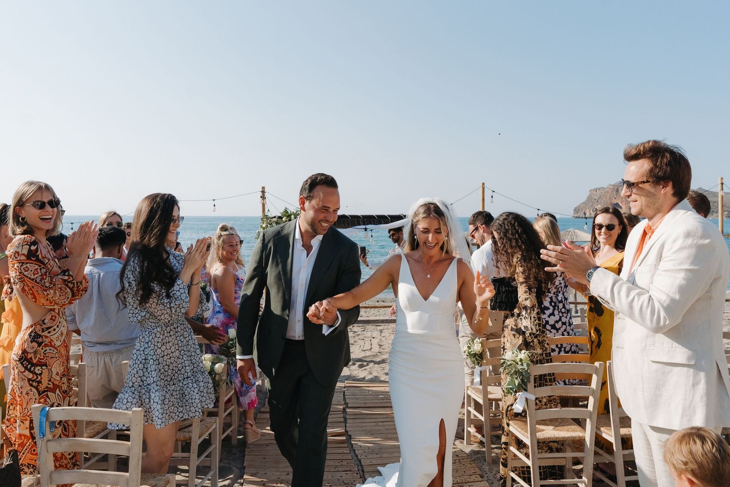 Wedding guests celebrating on a seaside deck with white wooden chairs and natural sunlight.