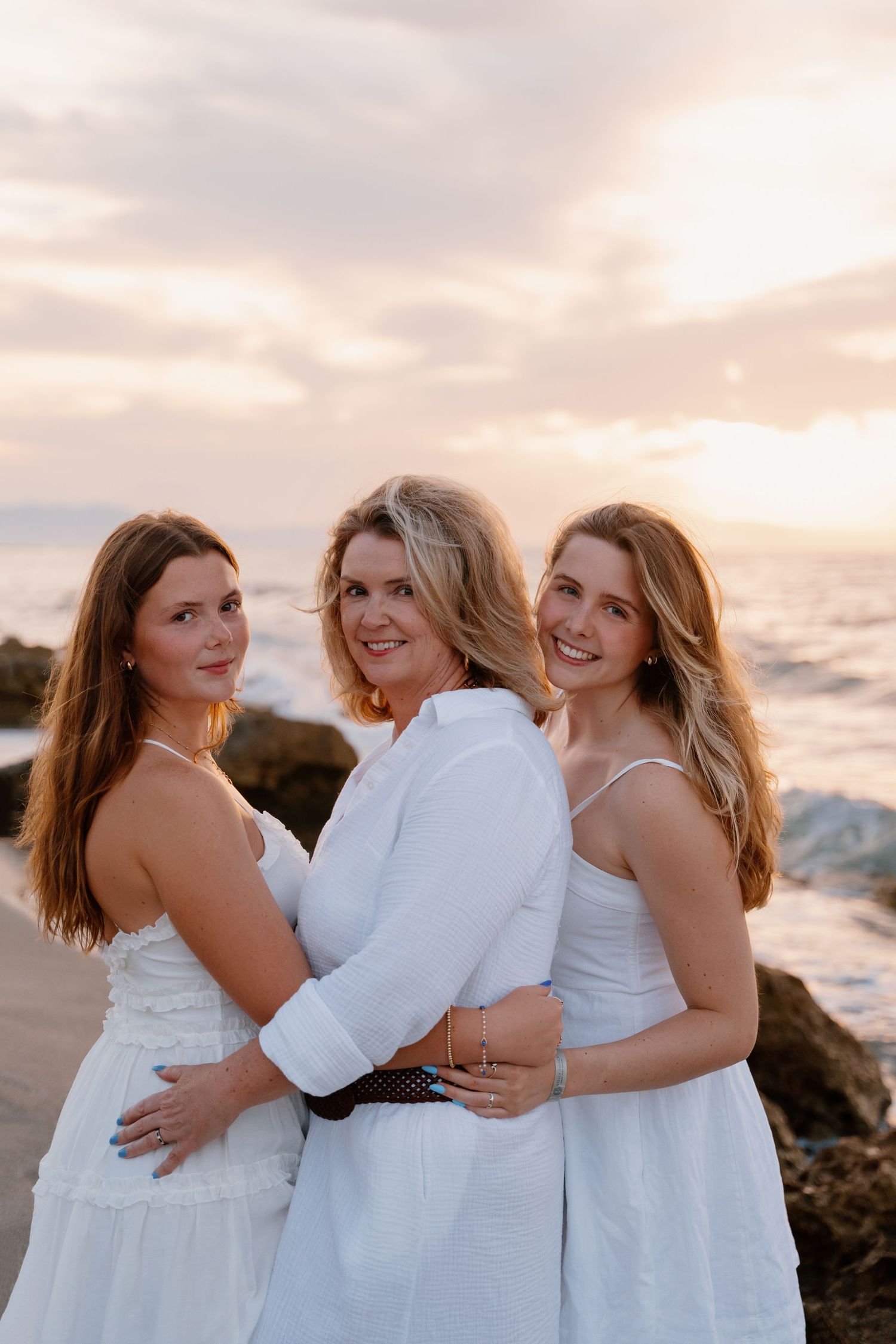 Three women in white dresses pose together on rocky beach at sunset.