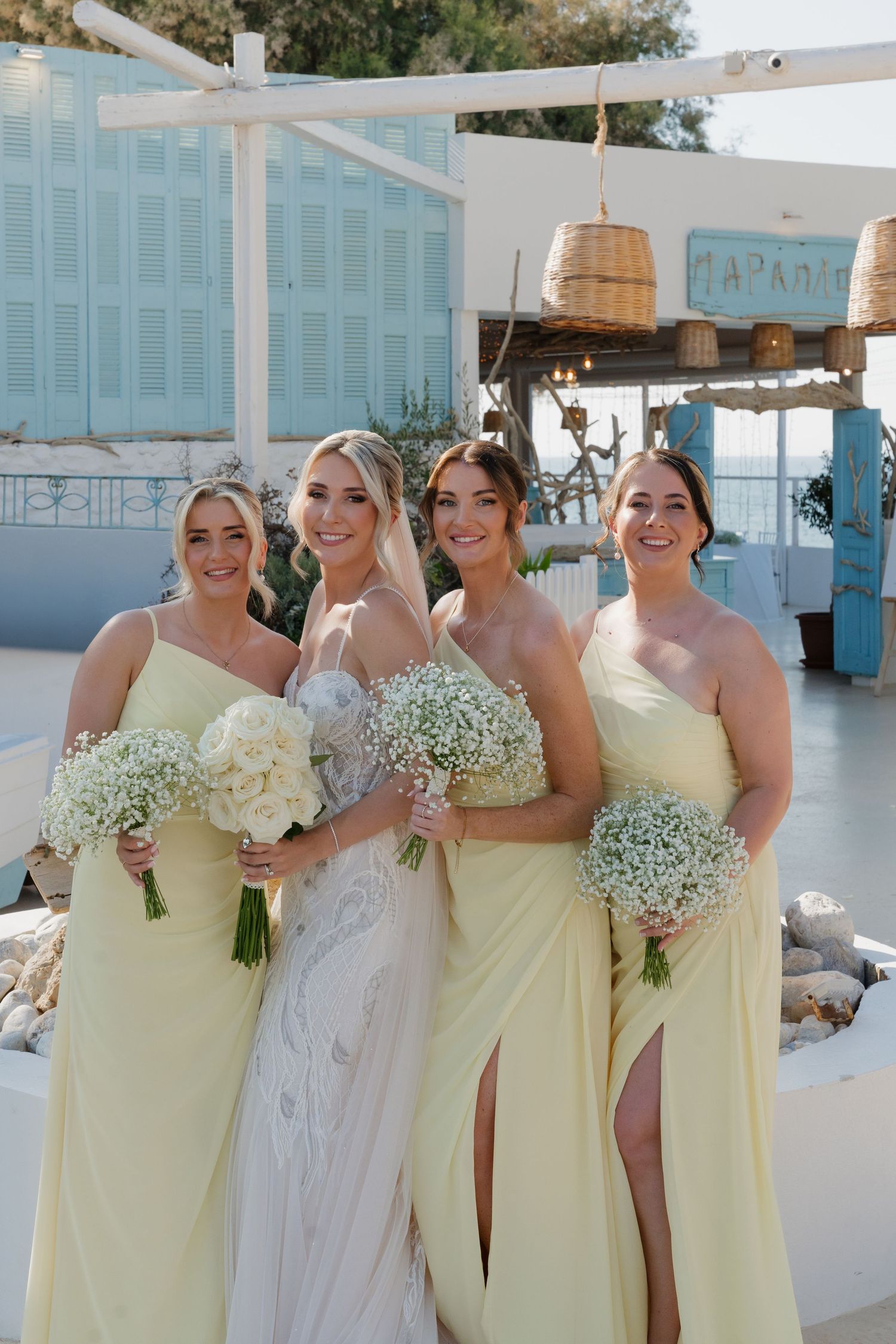 Bridesmaids in pale yellow dresses pose with bouquets at a coastal wedding venue with blue decor.