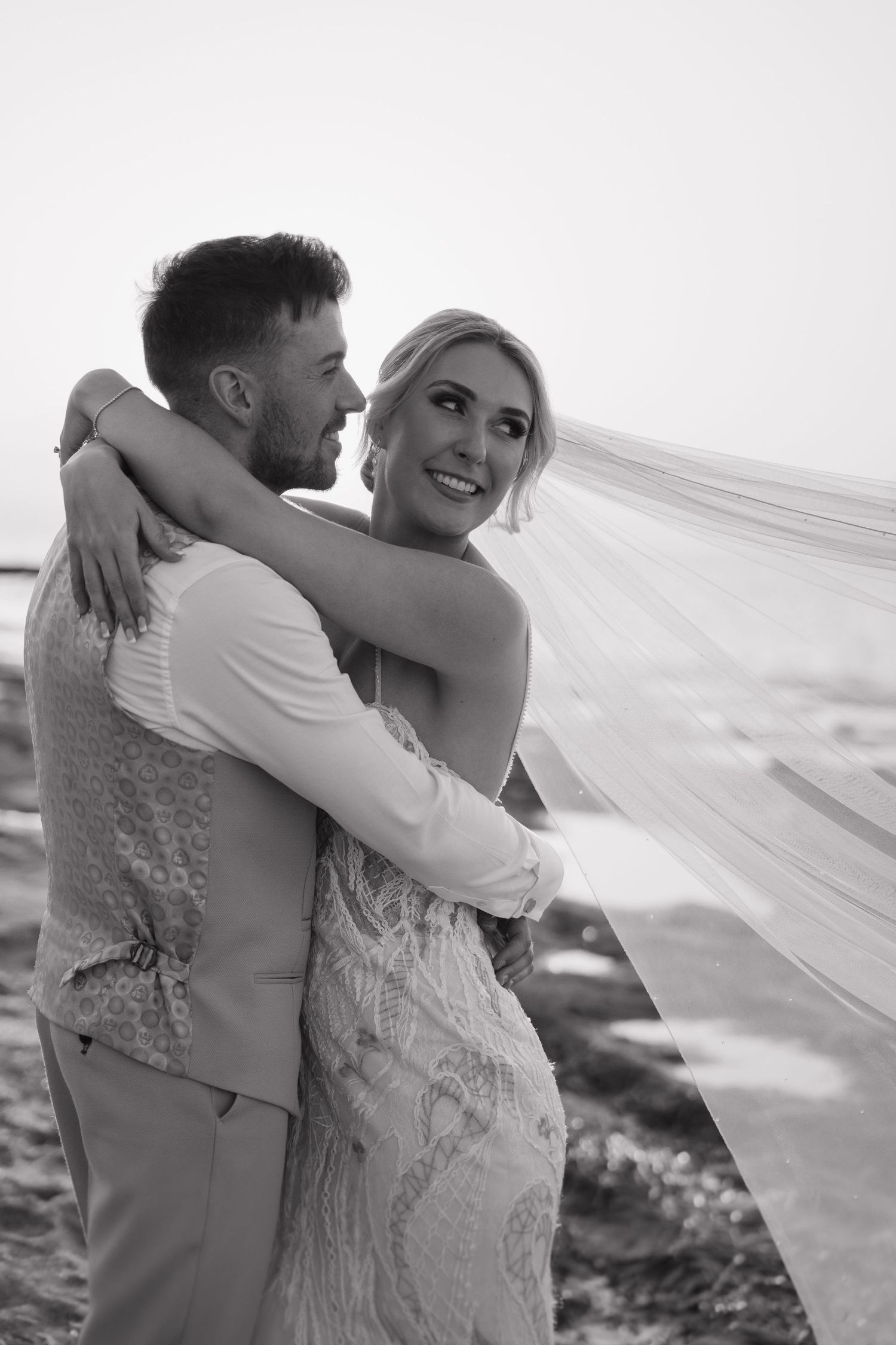 Black and white photo of happy couple embracing on beach with flowing veil.
