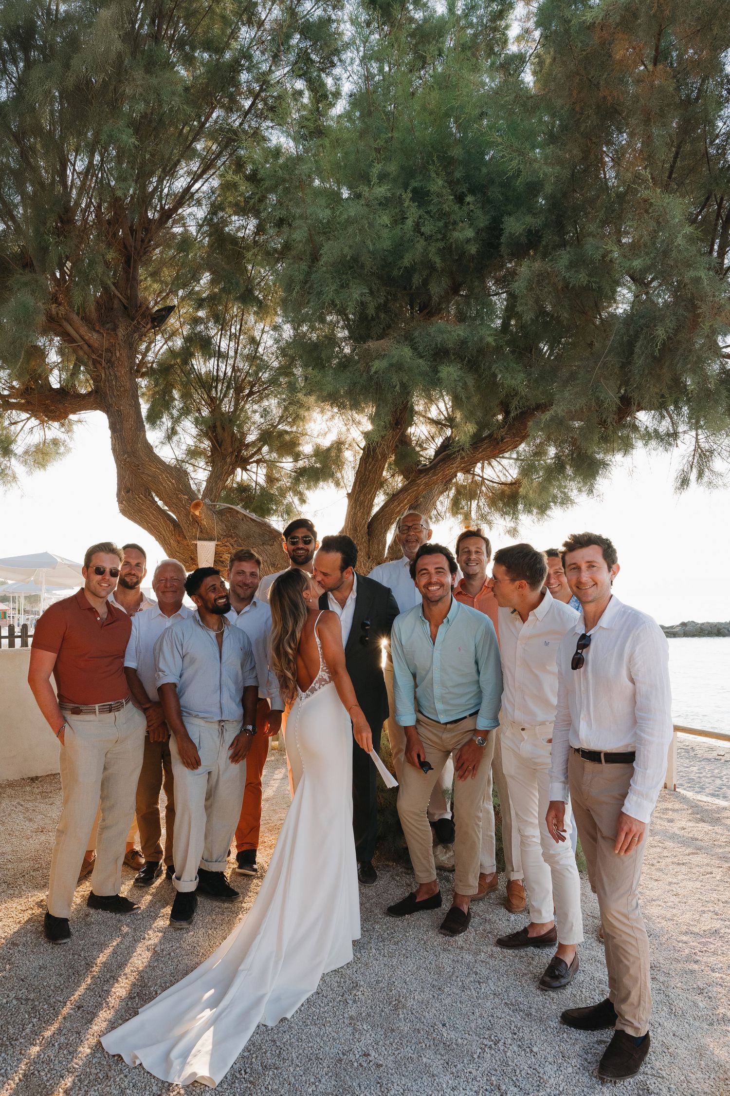 Wedding party poses together on a Mediterranean seaside terrace under a large pine tree during sunset.