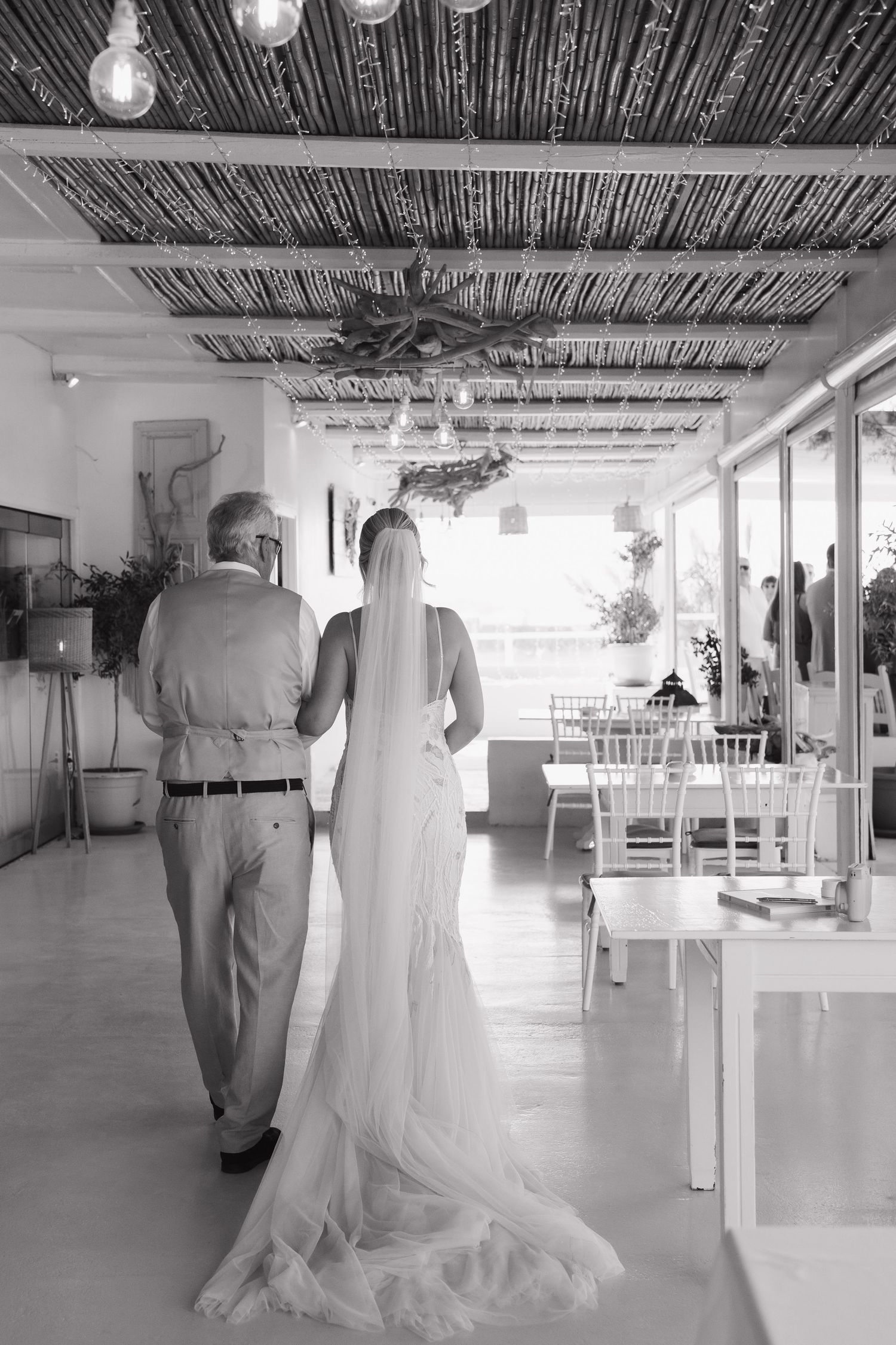 Black and white photo of a bride in flowing dress walking with companion through reception venue.