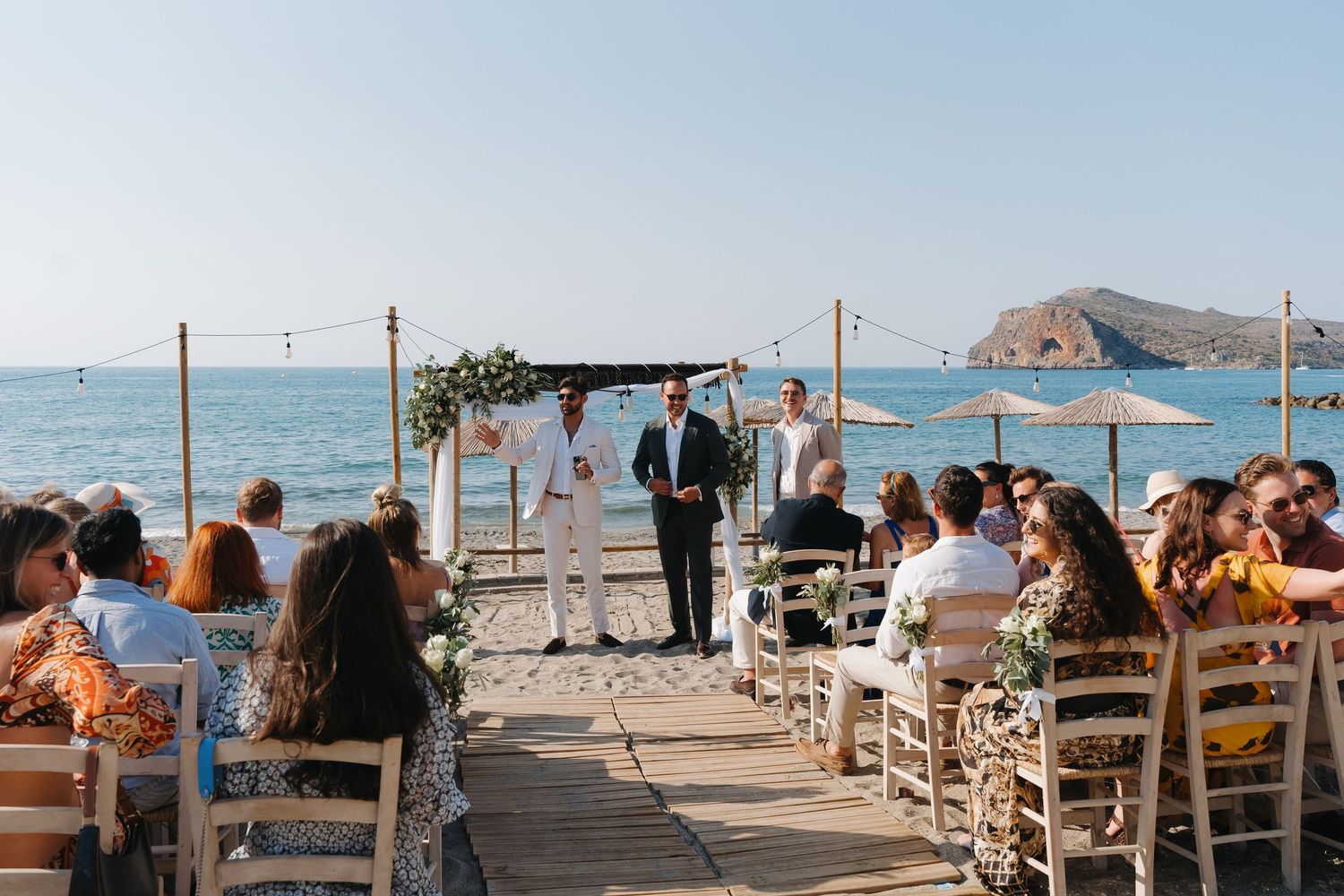 Beachfront wedding ceremony setup with wooden chairs and fabric canopy overlooking an ocean view and mountain landscape.