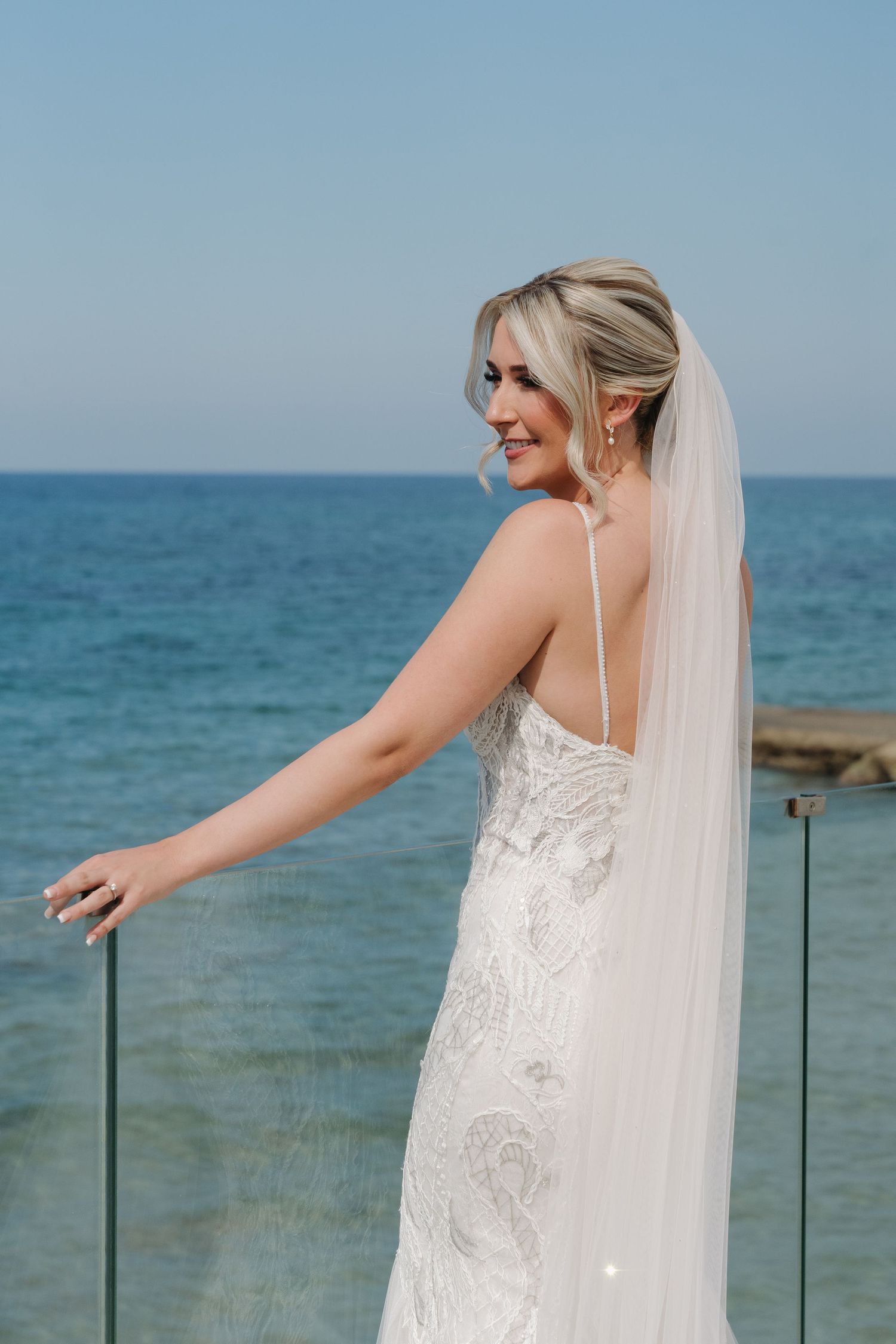 A person in a lace wedding dress and veil stands at an oceanfront balcony smiling.